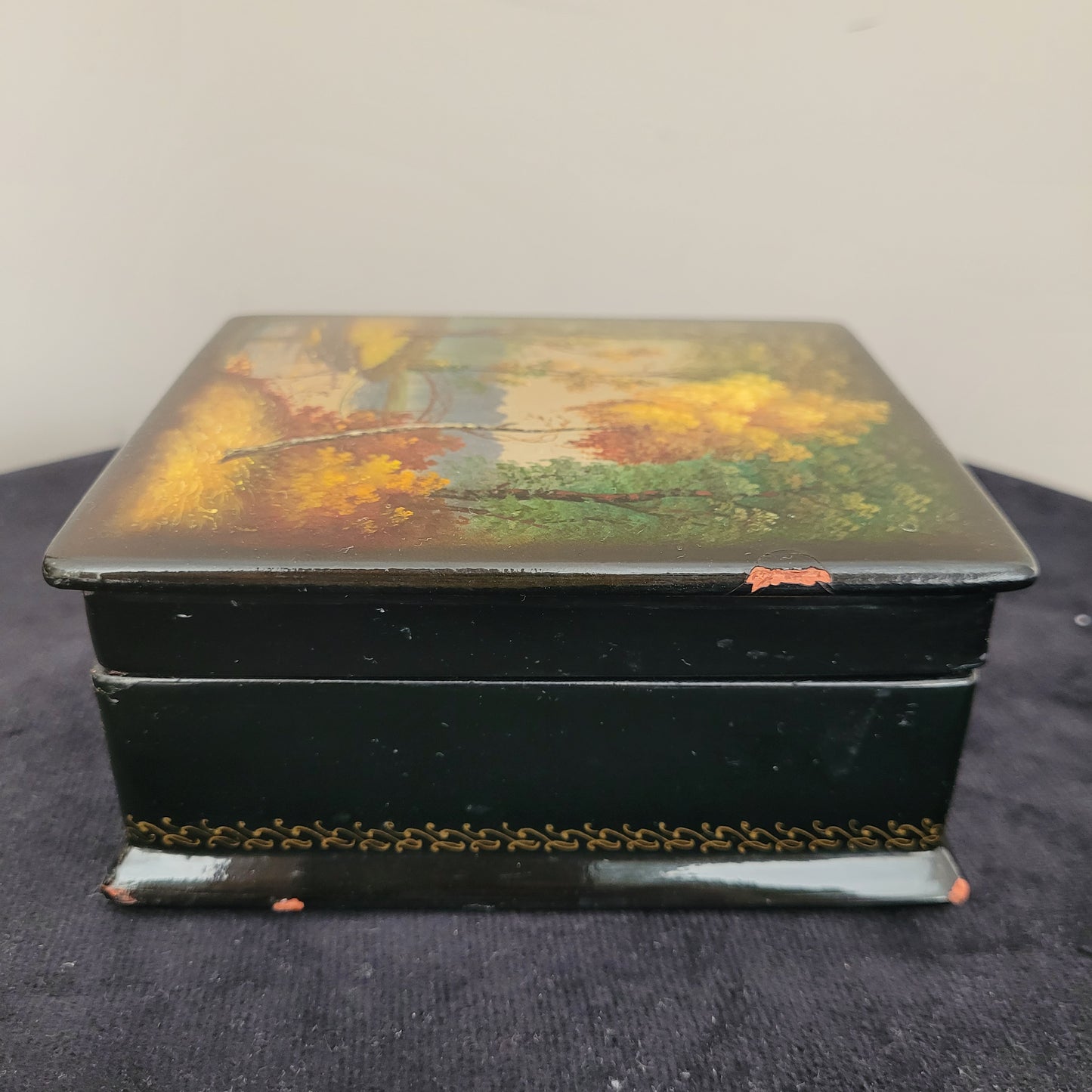Antike/Vintage Lackdose aus Holz aus der UdSSR – Seitenansicht mit kunstvollem Landschaftsmotiv Das Bild zeigt die Seitenansicht einer antiken/Vintage Lackdose aus Holz aus der UdSSR, verziert mit einer handgemalten, detailreichen Herbstlandschaft. Farbige Bäume in warmen Gelb-, Orange- und Grüntönen prägen das Motiv, während im Hintergrund eine elegante Brücke über einen Fluss oder See zu sehen ist. Der Deckel der Lackdose ist leicht geöffnet.