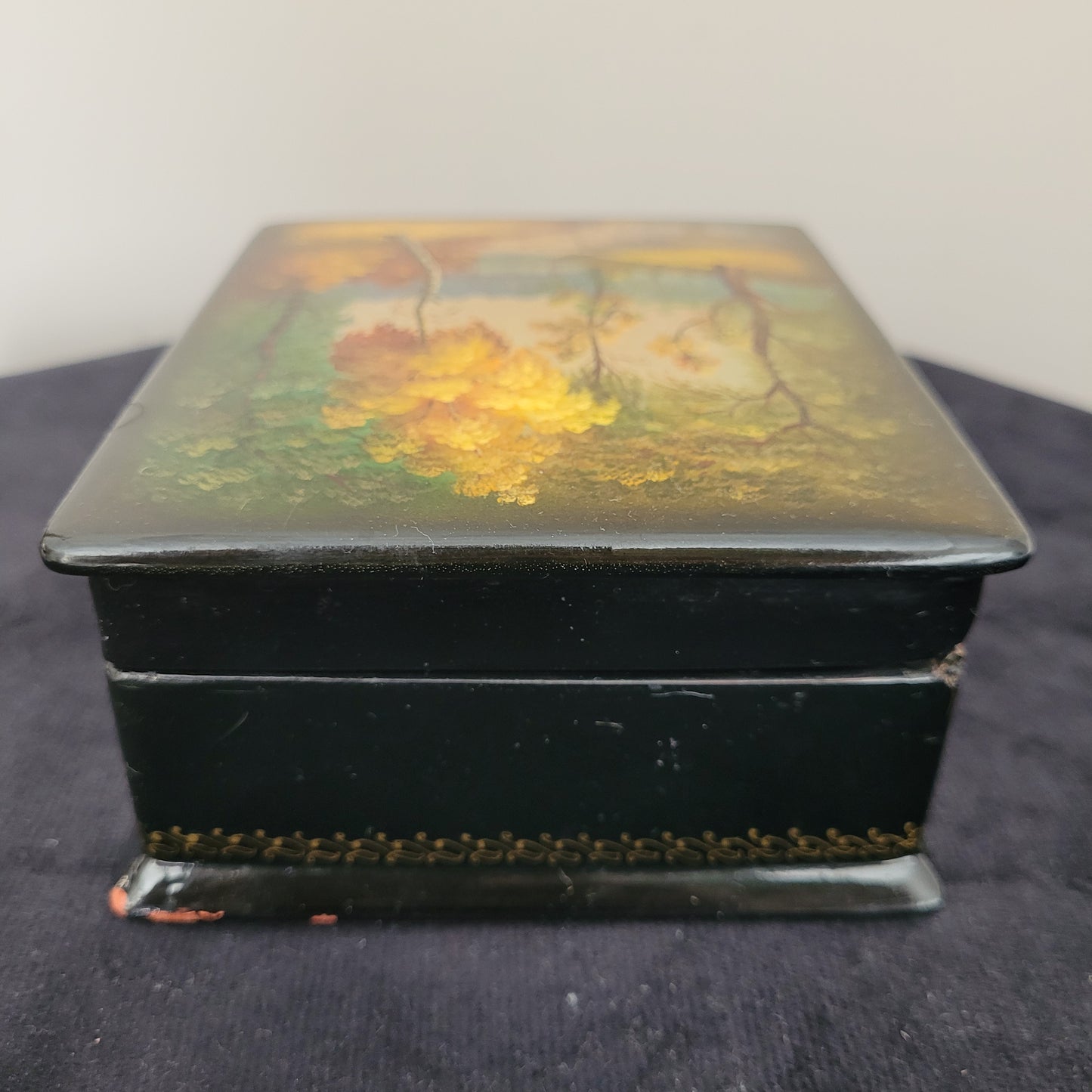 Antike/Vintage Lackdose aus Holz aus der UdSSR – Seitenansicht mit kunstvollem Landschaftsmotiv Das Bild zeigt die Seitenansicht einer antiken/Vintage Lackdose aus Holz aus der UdSSR, verziert mit einer handgemalten, detailreichen Herbstlandschaft. Farbige Bäume in warmen Gelb-, Orange- und Grüntönen prägen das Motiv, während im Hintergrund eine elegante Brücke über einen Fluss oder See zu sehen ist. Der Deckel der Lackdose ist leicht geöffnet.