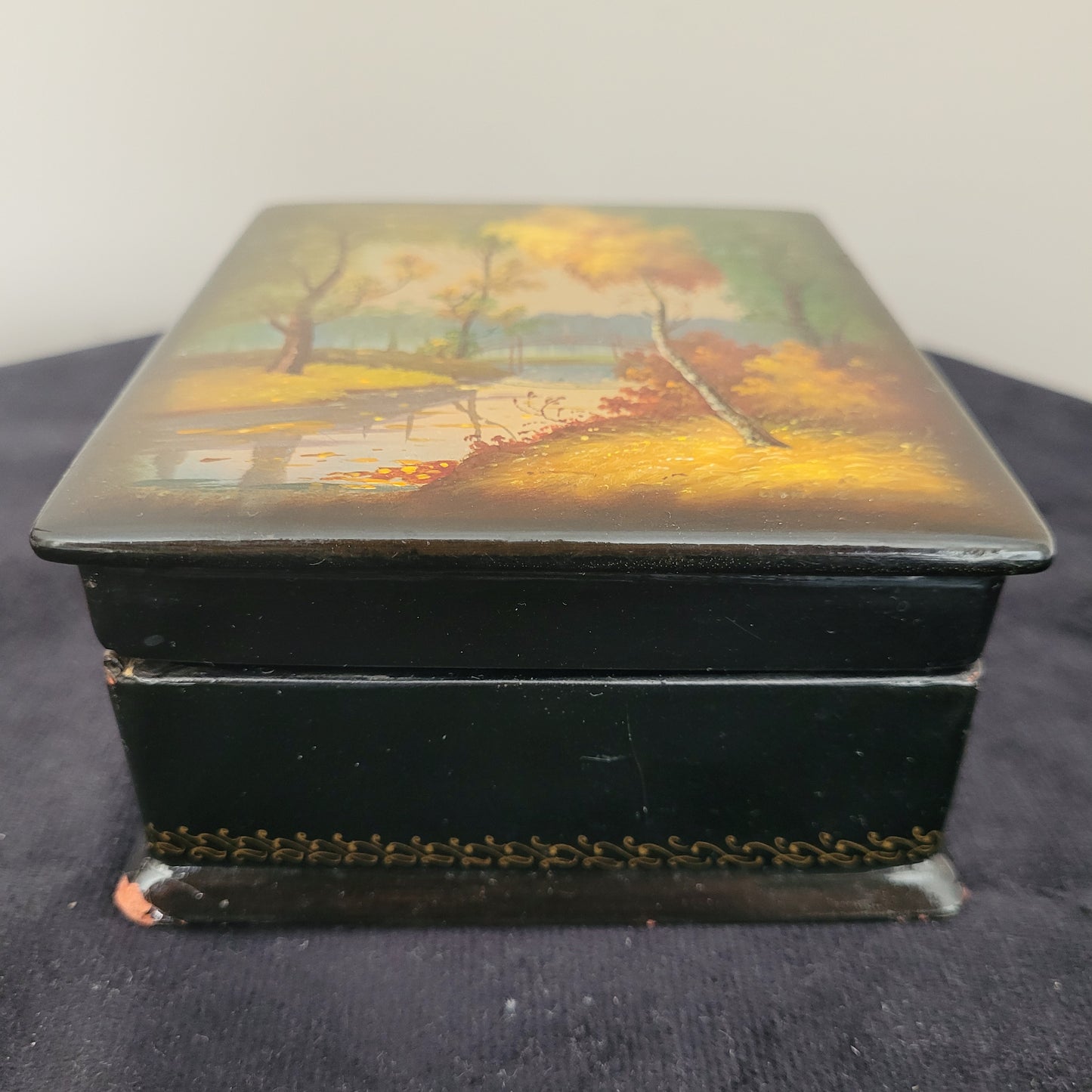 Antike/Vintage Lackdose aus Holz aus der UdSSR – Seitenansicht mit kunstvollem Landschaftsmotiv Das Bild zeigt die Seitenansicht einer antiken/Vintage Lackdose aus Holz aus der UdSSR, verziert mit einer handgemalten, detailreichen Herbstlandschaft. Farbige Bäume in warmen Gelb-, Orange- und Grüntönen prägen das Motiv, während im Hintergrund eine elegante Brücke über einen Fluss oder See zu sehen ist. Der Deckel der Lackdose ist leicht geöffnet.