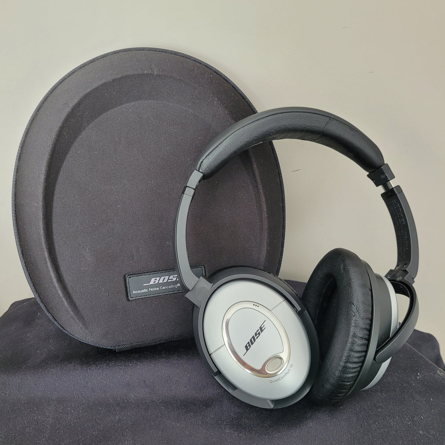 Die Bose Kopfhörer QuietComfort 15 liegen auf einer schwarzen, ovalen Tragetasche und präsentieren sich im klassischen Vintage-Design. Die silbernen Ohrmuscheln mit schwarzen Polstern unterstreichen den hochwertigen Look, während das Bose-Logo und die Modellbezeichnung „QuietComfort 15“ gut sichtbar sind. Die Tragetasche mit Bose-Branding vervollständigt das Set – ein Must-have für Liebhaber von Vintage-Elektronik und erstklassigen Kopfhörern.