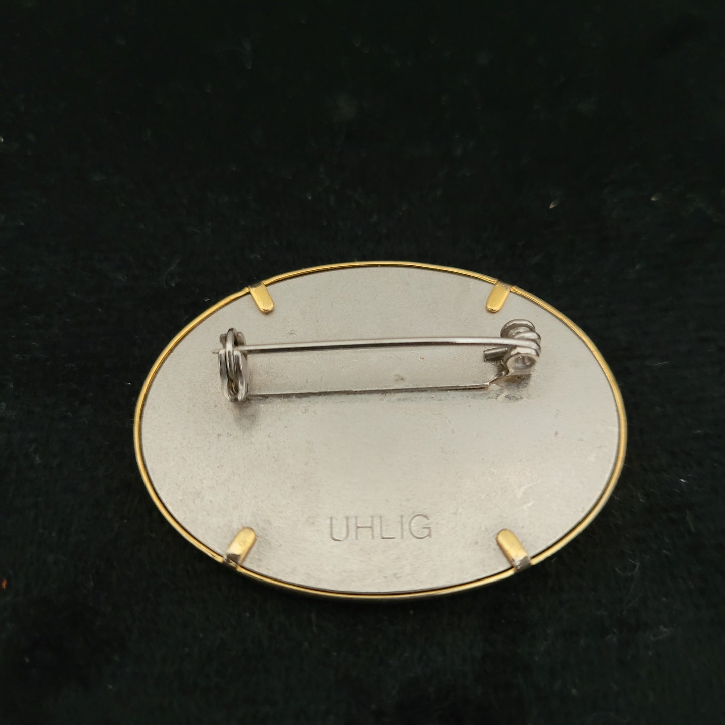 Rückseite einer ovalen Brosche mit Sicherheitsverschluss und Gravur 'UHLIG' auf der Metallplatte, befestigt durch vier goldene Klammern auf schwarzem Hintergrund.