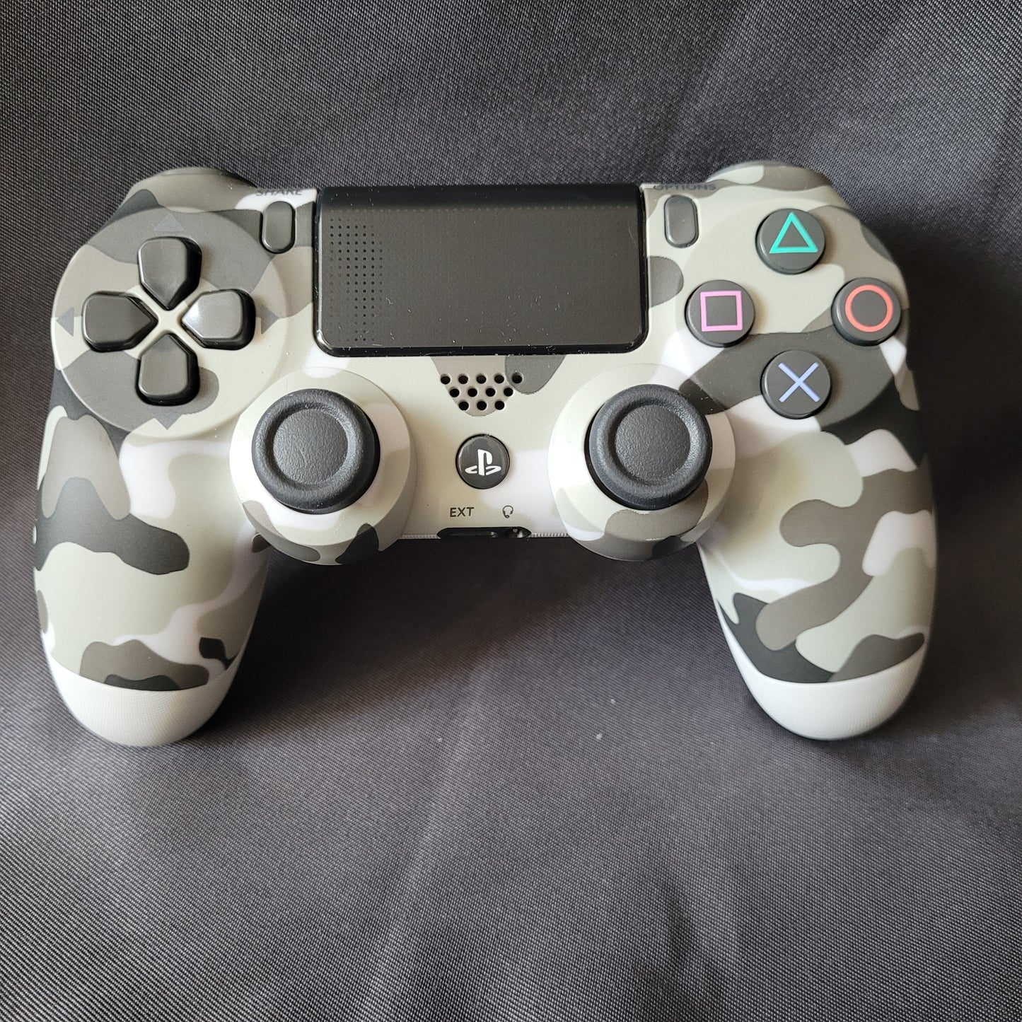 Original Sony Controller "Camouflage" – PlayStation 4 / PS4 – DualShock 4 in grau‑schwarz‑weißem Tarnmuster, mit Touchpad, Analog‑Sticks, Steuerkreuz und Aktionstasten, liegend auf dunkler Stoffoberfläche. EAN 0711719453512 / Modell CUH-ZCT1E.