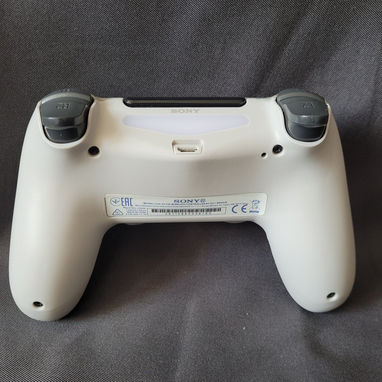 Rückseite eines Controller "Camouflage" – PlayStation 4 / PS4 – DualShock 4 – Original Sony in grau‑schwarz‑weißem Tarnmuster, mit sichtbaren L1‑ und R1‑Schultertasten, Lightbar, Micro‑USB‑Ladeanschluss und Modellaufkleber, liegend auf dunkler Stoffoberfläche. EAN 0711719453512 / Modell CUH-ZCT1E