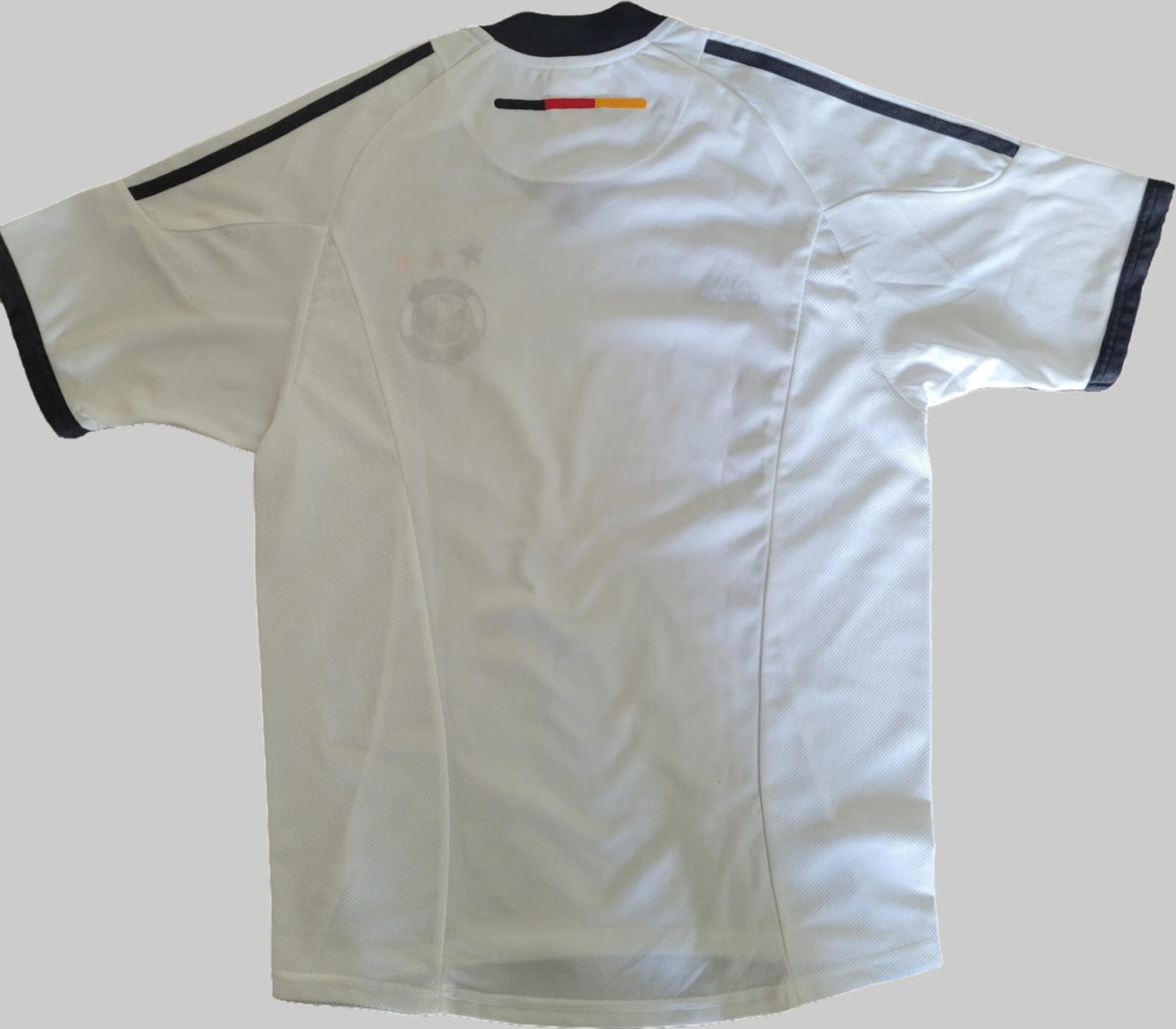 Deutschland Vintage Trikot 2002 – Rückseite auf grauem Hintergrund Das Bild zeigt die Rückseite eines Deutschland Vintage Trikots aus der WM 2002, platziert auf einem grauen Hintergrund. Das Trikot ist weiß mit schwarzen Akzenten an Kragen und Ärmeln. Die Rückseite ist schlicht gehalten, ohne sichtbare Rückennummer oder Schriftzüge