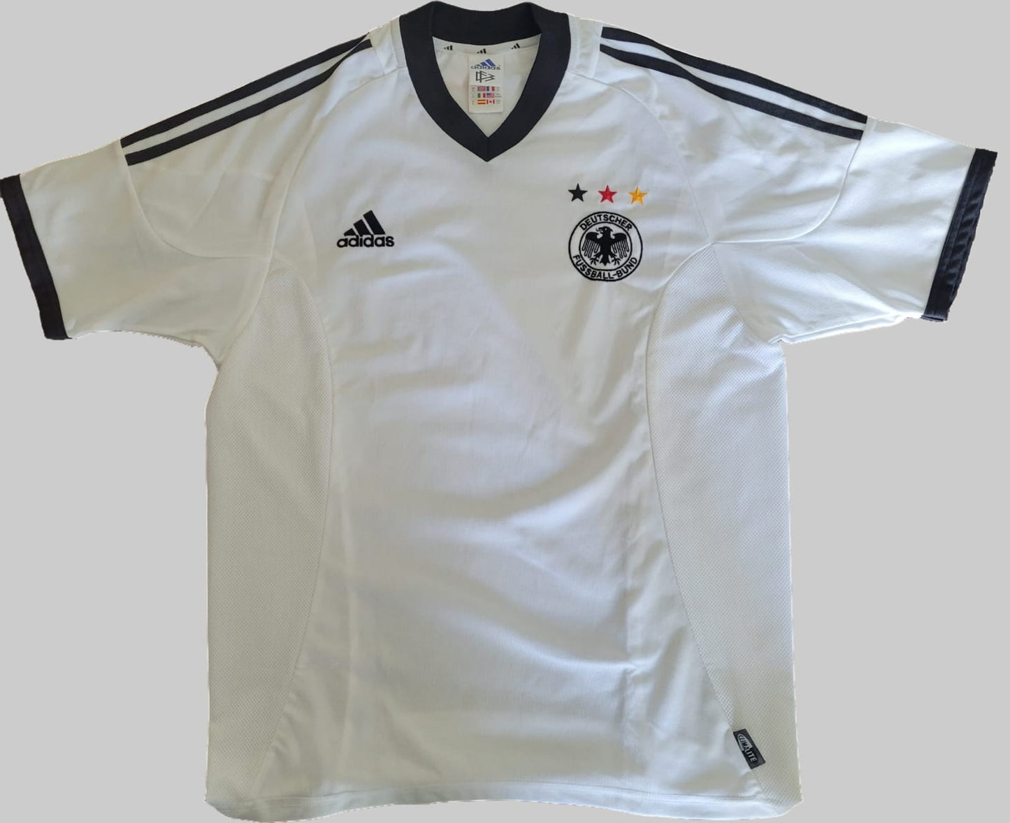 Deutschland Vintage Trikot 2002 – Vorderseite auf grauem Hintergrund Das Bild zeigt die Vorderseite eines Deutschland Vintage Trikots aus der WM 2002, platziert auf einem grauen Hintergrund. Das Trikot ist weiß mit schwarzen Akzenten an den Ärmeln und Seiten. Auf der linken Brustseite befindet sich das DFB-Logo mit den deutschen Nationalfarben, während auf der rechten Brustseite das Adidas-Logo sichtbar ist. In der Mitte prangt das Sponsorenlogo in dunkler Schrift. Das Trikot besitzt einen schwarzen Kragen