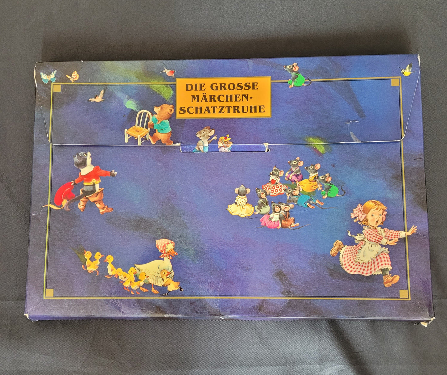 Vintage Kinderbuch-Set „Die große Märchenschatztruhe“ mit farbenfroher Illustrationen klassischer Märchenfiguren auf dunkelblauem Hintergrund. Die nostalgische Pappbox zeigt detailreiche Szenen mit Tieren in Kleidung, märchenhaften Charakteren und liebevoll gestalteten Motiven – ideal für Sammler und Liebhaber von Vintage-Märchenbüchern.