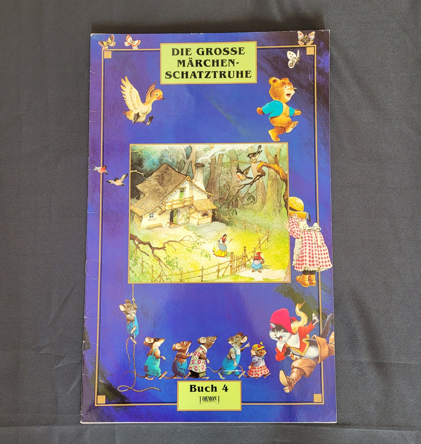 Vintage Cover des Märchenbuchs „Die große Märchenschatztruhe – Buch 4“ mit detailreichen Illustrationen klassischer Märchenfiguren wie Rotkäppchen, sprechenden Mäusen und einem Waldhaus auf tiefblauem Hintergrund mit Sternen. Das nostalgische Design und die liebevoll gestalteten Szenen machen dieses Vintage-Märchenbuch zu einem begehrten Sammlerstück für Märchenliebhaber und Fans traditioneller Kinderliteratur.