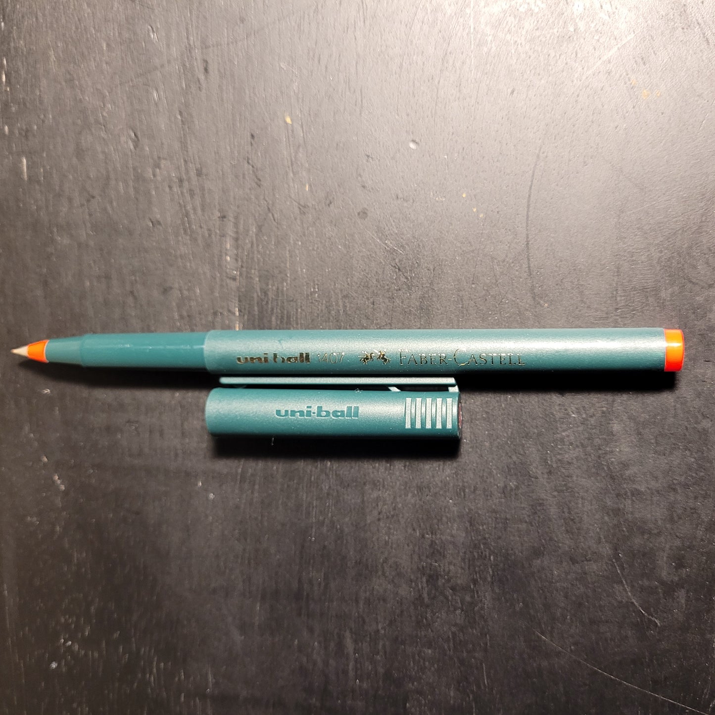 Ein grüner Faber-Castell Stift mit einer orangefarbenen Spitze und einer abgenommenen Kappe. Der Stift trägt die Aufschriften 'uni-ball 407' und 'FABER-CASTELL'. Die Kappe liegt neben dem Stift und trägt ebenfalls die Aufschrift 'uni-ball'. Der Stift liegt auf einer dunklen Oberfläche, möglicherweise ein Schreibtisch oder eine Tischplatte.