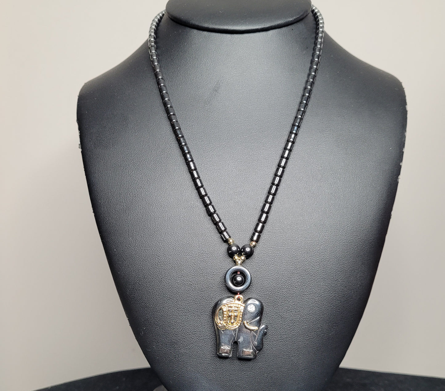 Ein einzigartiges Halsband aus schwarzen Perlen, präsentiert auf einem Schmuckständer. Der detaillierte Elefanten-Anhänger ist mit goldenen Akzenten und kleinen Perlen verziert.