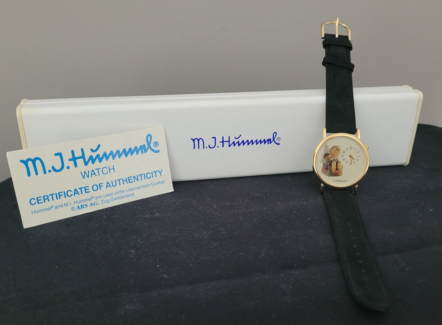 Das Bild zeigt die Vorderseite einer M.J. Hummel Armbanduhr mit einem eleganten, goldfarbenen Gehäuse und einem schwarzen Lederarmband. Im Zentrum stechen das kindlich illustrierte Zifferblatt und das daneben platzierte Zertifikat der Authentizität hervor, auf dem unter anderem die Unterschriften „M.J. Hummel WATCH CERTIFICATE OF AUTHENTICITY“ und Lizenzinformationen stehen. Im Hintergrund befindet sich eine weiße Box mit dem blauen M.J. Hummel Logo.