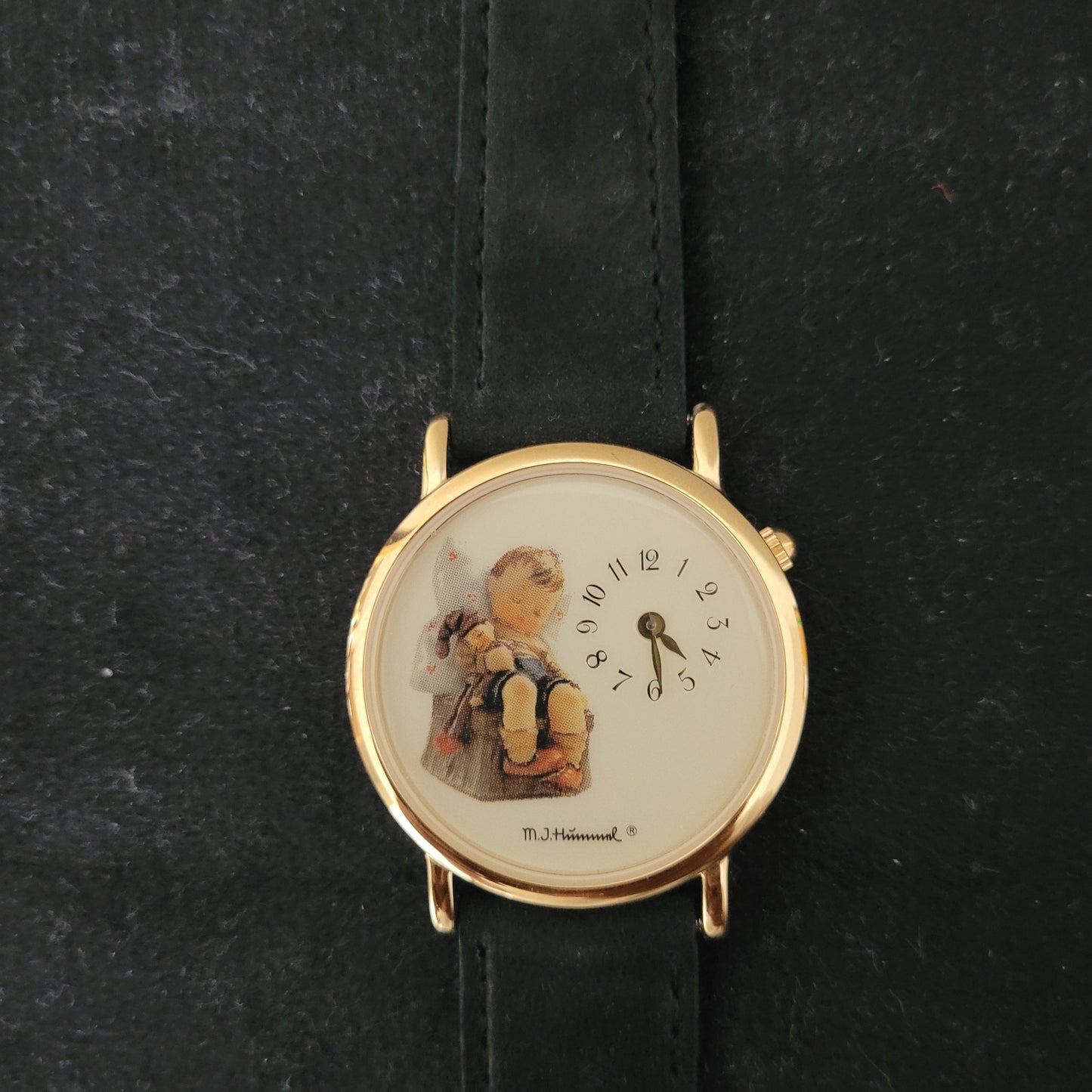 Das Bild zeigt die Vorderseite einer M.J. Hummel Vintage Armbanduhr mit einem eleganten, goldfarbenen Gehäuse und einem schwarzen Lederarmband. Das kunstvoll gestaltete Zifferblatt ist mit einer Illustration eines Kindes, das eine Puppe hält, versehen, und rechts am Zifferblatt ist der Schriftzug „M.J. Hummel“ erkennbar – ein authentisches Original-Vintage-Design.