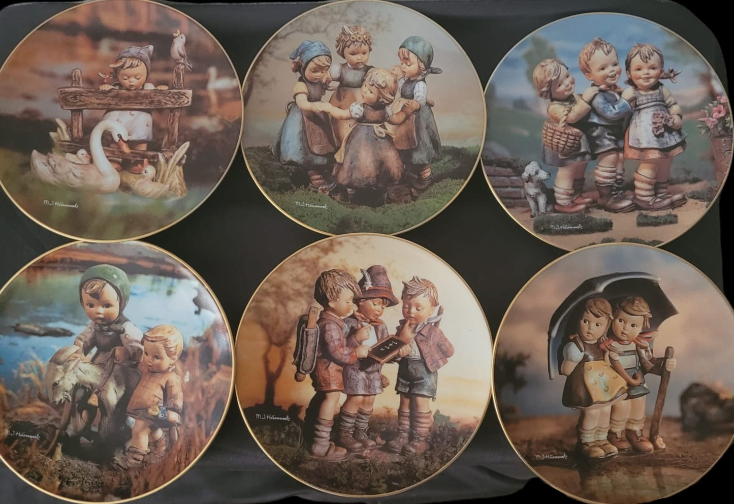 Sechs dekorative M.J. Hummel Sammelteller, jeweils mit liebevoll gestalteten Kinderszenen, darunter spielende Kinder, Tiere und ländliche Hintergründe; die Teller sind in zwei Reihen zu je drei Stück angeordnet und zeigen detailreiche Motive mit warmen Farben.