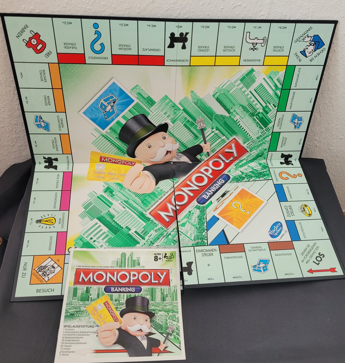 Das Bild zeigt das Spielbrett des Monopoly Banking Gesellschaftsspiels von Hasbro sowie die dazugehörige Spielanleitung. Das Spielbrett präsentiert die klassischen Monopoly-Elemente, ergänzt durch moderne Banking-Technologie mit einem elektronischen Kartenlesegerät. Die Spielanleitung liegt neben dem Brett und bietet eine klare Übersicht über die Regeln und Spielmechaniken. Das Bild wird gerade erstellt!