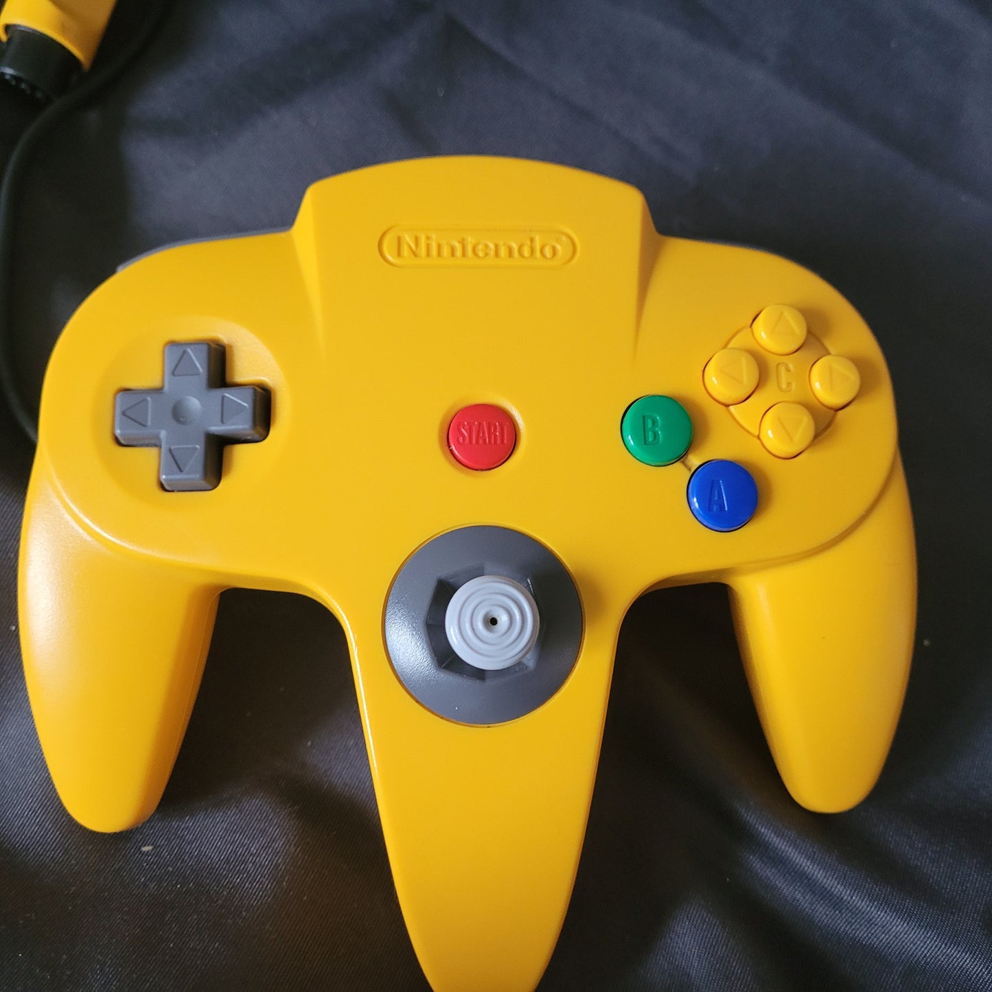 Nintendo 64 – N64 – Gelber Controller/Joystick Model NUS-005A auf dunklem Stoffhintergrund. Der Controller zeigt das klassische dreizackige Design mit grauem Steuerkreuz links, grauem Analogstick und rotem Startknopf in der Mitte sowie bunten Tasten rechts (vier gelbe C-Tasten, grüner B-Button, blauer A-Button). Oben ist das Nintendo-Logo eingeprägt, das Kabel mit Stecker ist sichtbar. Retro/Vintage Zubehör für die N64 Konsole.