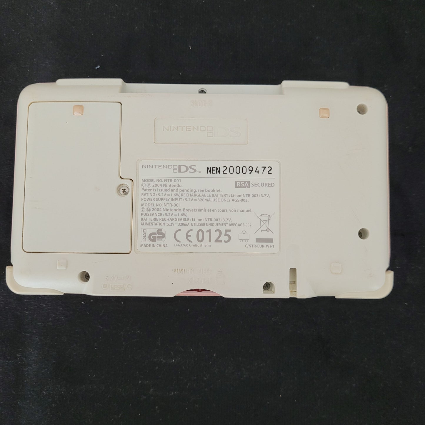 Unterseite der Nintendo DS Pink Model No NTR-001 Handheld Konsole, liegend auf schwarzem Stoff. Die Rückseite zeigt das Batteriefach, die Modellnummer und die Steckplätze für Nintendo DS Game Cards sowie Game Boy Advance Cartridges. Gebrauchsspuren sind sichtbar, die Konsole bleibt jedoch voll funktionsfähig – ein perfektes Retro-Sammlerstück für Gaming-Fans.