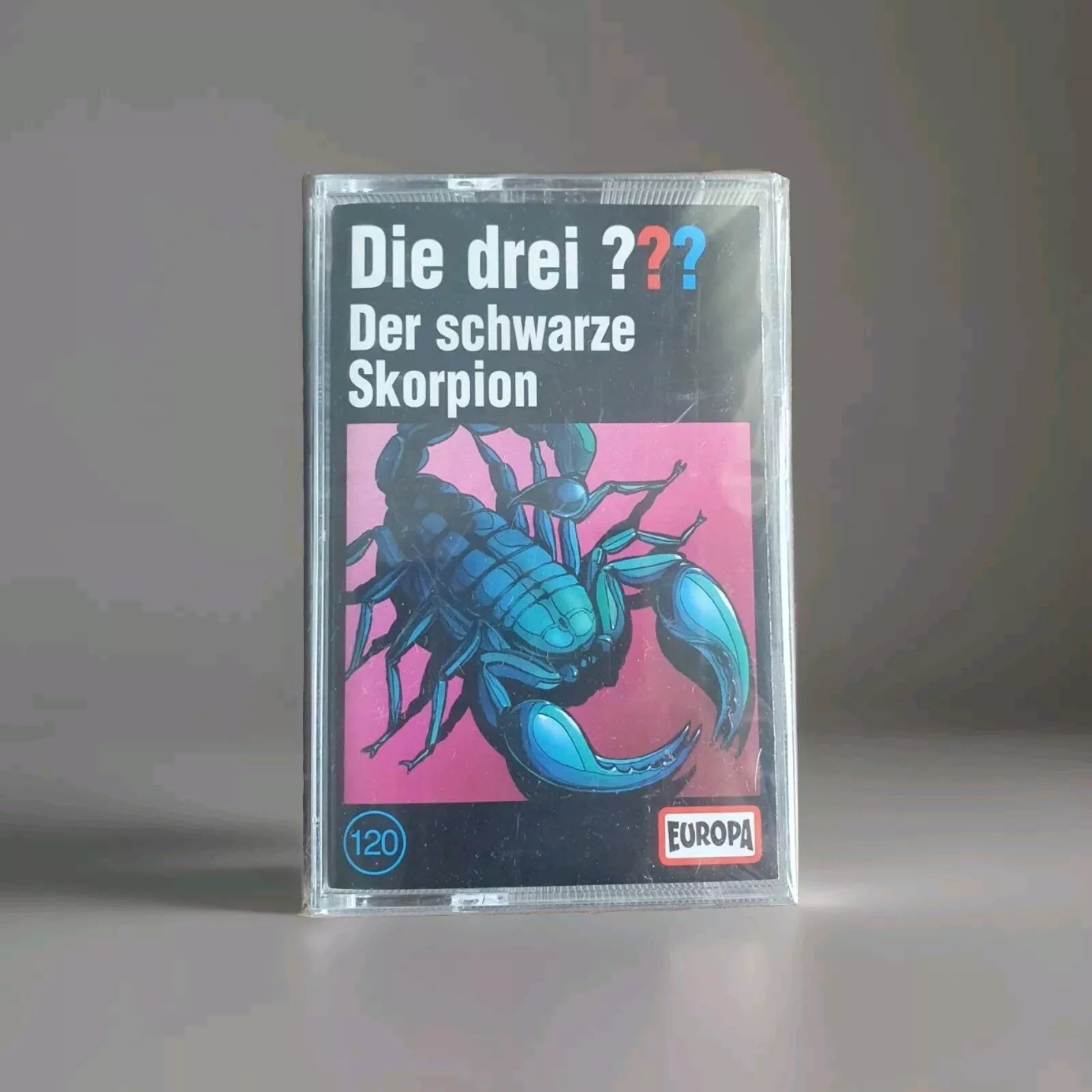 „Kassette 'Die drei ??? Der schwarze Skorpion' - Illustration eines blauen Skorpions auf rosa Hintergrund. Hörspielkassette Nr. 120 vom Label 'EUROPA'.“