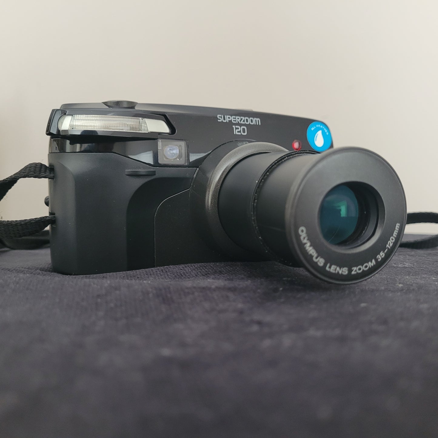 Olympus Superzoom 120 Multi AF Analogkamera – klassische Vintage Analogkamera mit ausgefahrenem 35-120mm Olympus Zoomobjektiv. Die elegante schwarze Kamera liegt auf dunklem Stoff und zeigt deutlich die Modellbezeichnung Superzoom 120. Perfekt für Liebhaber hochwertiger Retro-Fotografie.