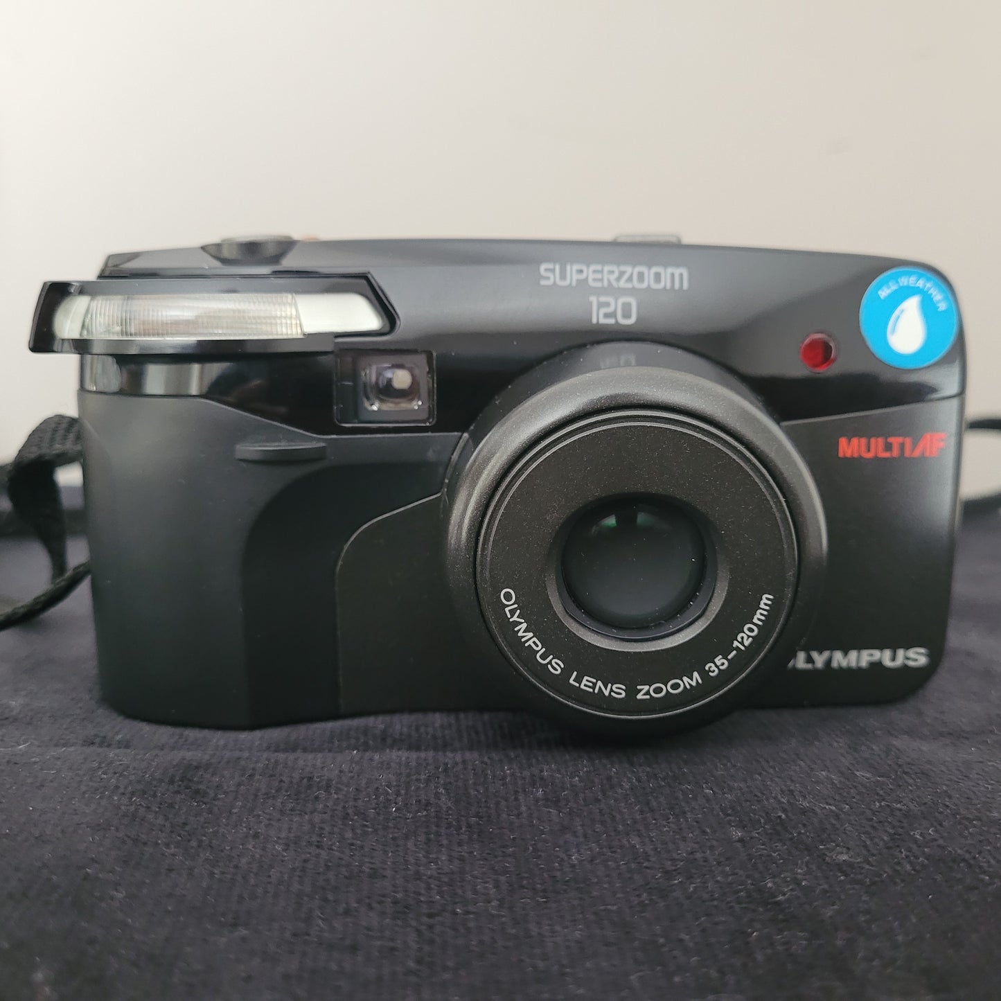 Vorderansicht der Olympus Superzoom 120 Multi AF Analogkamera, mit geschlossenem Linsendeckel. Die Modellbezeichnung Superzoom 120 und die Zoomangabe Olympus Lens Zoom 35-120mm sind gut sichtbar. Die elegante schwarze Vintage Analogkamera liegt auf dunklem Stoff, mit Tragegurt an der Seite – perfekt für Liebhaber klassischer Analogfotografie.