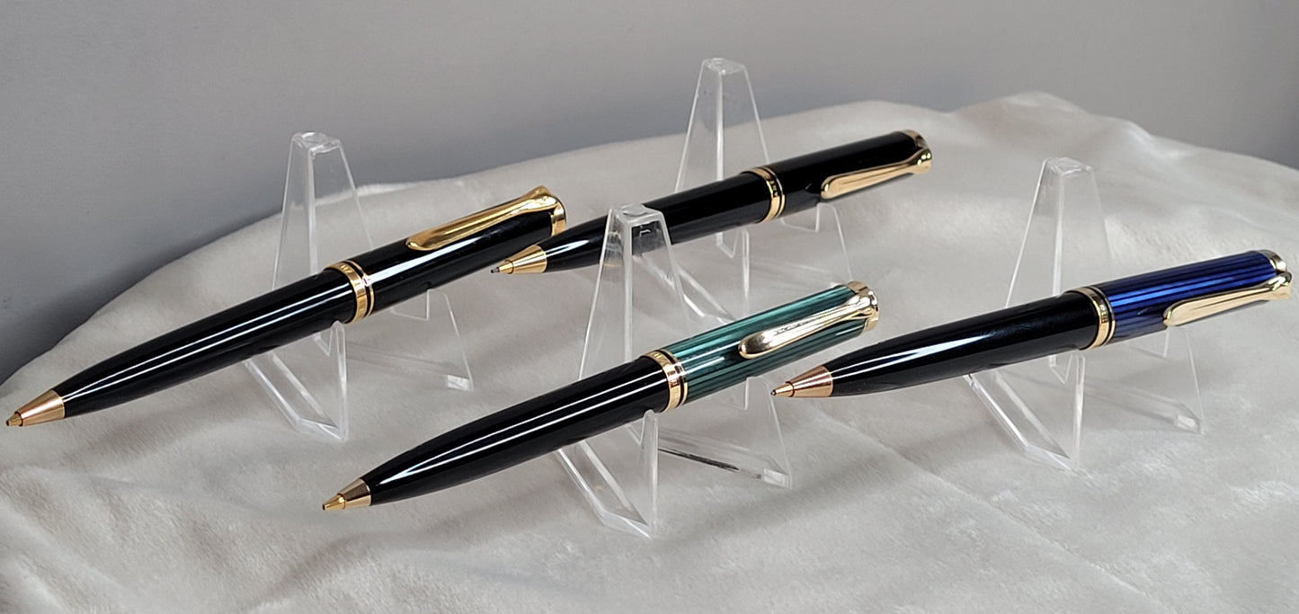 Vier hochwertige Pelikan Souverän Drehbleistifte in verschiedenen Modellen, präsentiert auf durchsichtigen Kunststoffständern. Die Stifte bestechen durch ihr elegantes Design in Schwarz, Blau und Grün, verziert mit goldenen Akzenten und verzierten Clips. Diese luxuriösen Schreibgeräte, hergestellt in Deutschland, stehen für höchste Verarbeitungsqualität und Präzision. Ideal als stilvolles Accessoire für das Büro oder als luxuriöses Geschenk.