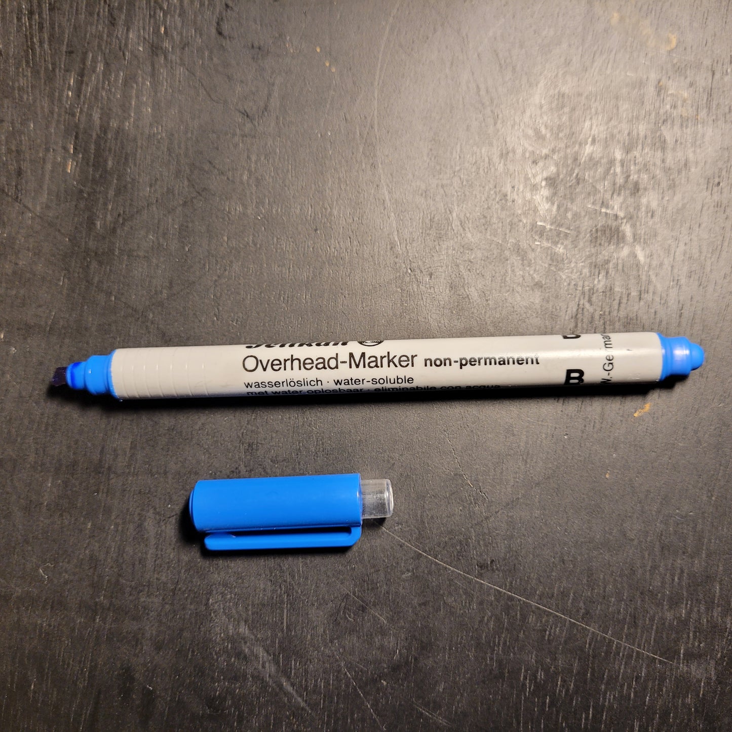 Die Abbildung zeigt einen Overhead-Marker mit abgenommener blauer Kappe, die daneben liegt. Der Marker ist mit der Aufschrift "Overhead-Marker non-permanent" versehen und als "wasserlöslich - water-soluble" beschrieben. Der Marker hat eine blaue Spitze und eine blaue Kappe, die unten neben dem Marker liegt. Der Marker liegt auf einer dunklen Oberfläche, möglicherweise einem Schreibtisch oder Tisch.