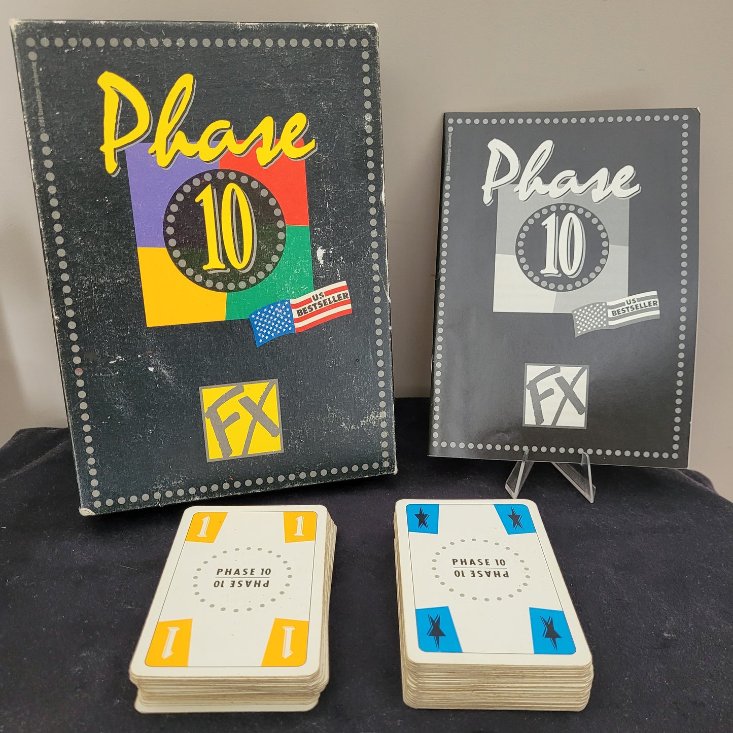 Das originale Phase 10 FX 1998 Ravensburger Vintage Version Gesellschaftsspiel aus Deutschland. Das Bild zeigt das vollständige Spielset mit zwei Stapeln Spielkarten und zwei Anleitungen – eine in Schwarz-Weiß, die andere farbig und mit Gebrauchsspuren. Dieses klassische Ravensburger Gesellschaftsspiel ist ein echtes Vintage-Highlight für Sammler und Liebhaber von Retro-Spielen.
