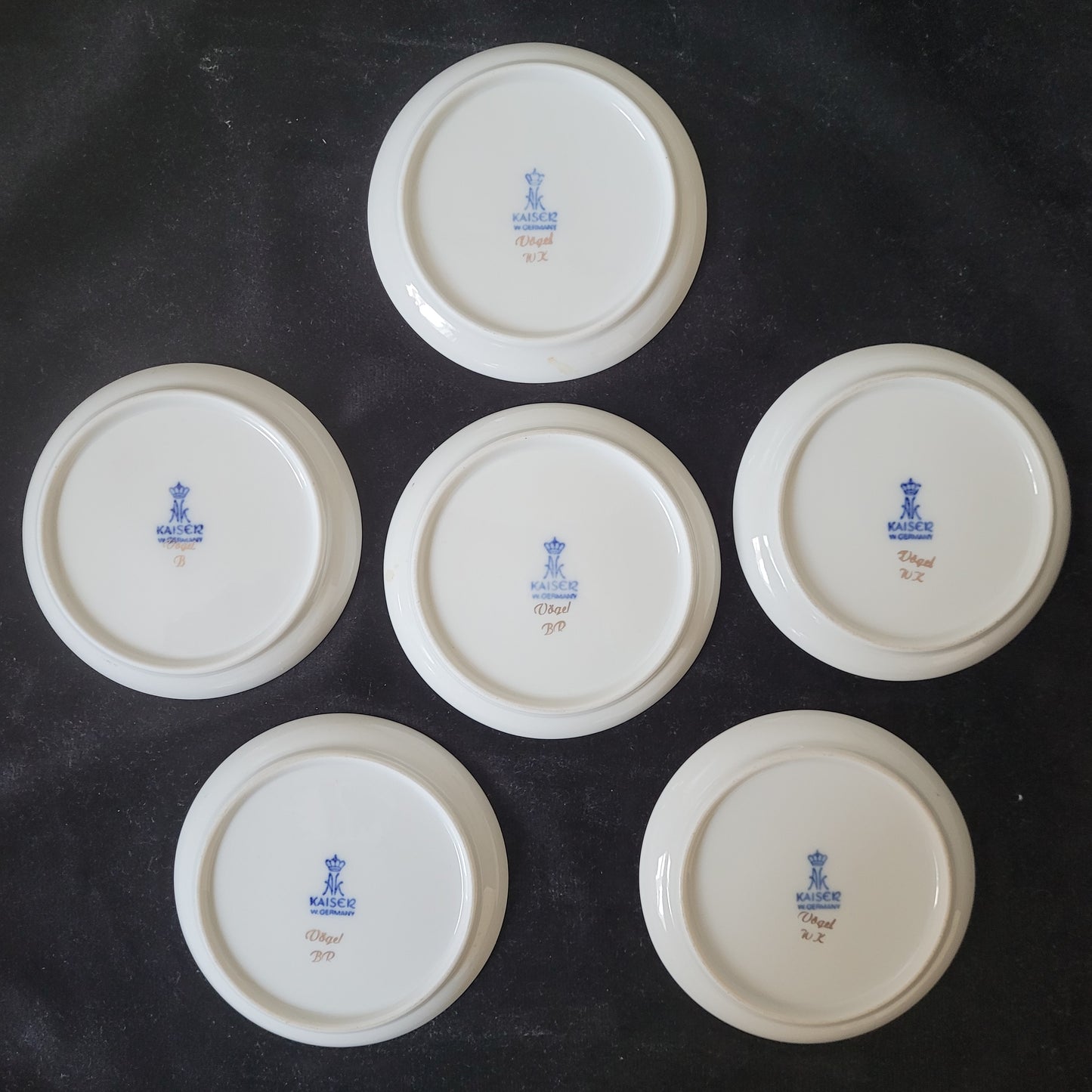 Die Rückseiten der 6-teiligen Porzellan Schalen / Teller - Manufaktur Kaiser - Vögel sind auf dunklem Hintergrund angeordnet. Sichtbar sind die Markierungen der Manufaktur Kaiser sowie die fein gearbeitete Porzellanstruktur. Die elegante Verarbeitung und das hochwertige Material unterstreichen den Vintage-Charme dieses antiken Sammlerstücks – ein Highlight für Liebhaber klassischer Porzellankunst.