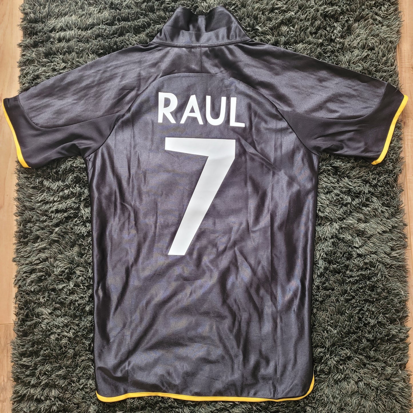 Rückansicht des Vintage Real Madrid Trikots Raul Saison 1999/2000 in Größe S, flach ausgelegt auf dunkelgrünem Teppich. Schwarzes Adidas-Trikot mit gelbem Saum zeigt den legendären Spielername "RAUL" und die Rückennummer 7 in Weiß – ein echtes Highlight für Sammler und Fans von Real Madrid Vintage Fußballtrikots