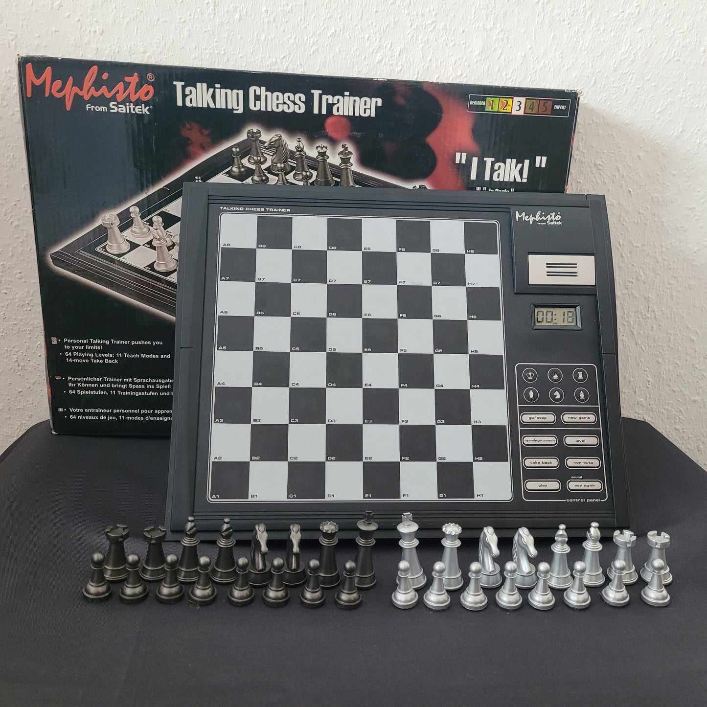 Das Bild zeigt das Schach Mephisto Talking Chess Trainer Modul von Saitek mit vollständigem Zubehör in seiner Originalverpackung (OVP). Das Schachgerät liegt auf einem schwarzen Untergrund mit weißem Hintergrund, wodurch die Details der Verpackung und des Moduls gut sichtbar sind. Neben dem Schachcomputer sind die Schachfiguren erkennbar, was die Vollständigkeit des Sets unterstreicht.