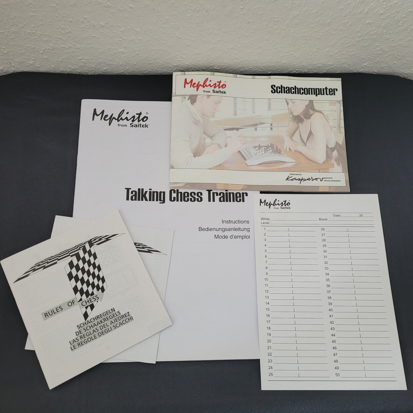 Das Bild zeigt die Betriebsanleitungen des Schach Mephisto Talking Chess Trainer von Saitek. Die Anleitungen liegen auf einem schwarzen Untergrund mit weißem Hintergrund und enthalten detaillierte technische Informationen zum Schachcomputer. Die einzelnen Seiten sind übersichtlich gestaltet und vermitteln wichtige Hinweise zur Bedienung und den Funktionen des Geräts.