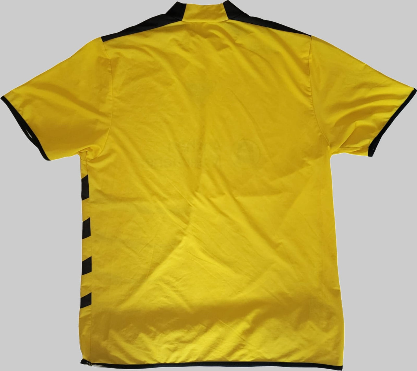 TSV Alemannia Aachen Vintage Trikot 2006/2007 – Rückseite auf grauem Hintergrund Das Bild zeigt die Rückseite des TSV Alemannia Aachen Vintage Trikots 2006/2007, platziert auf einem grauen Hintergrund. Die Rückseite ist schlicht gehalten, ohne sichtbare Rückennummer oder Signaturen. Das klassische Design mit gelber Grundfarbe und schwarzen Akzenten spiegelt die Vereinsfarben wider und unterstreicht den Vintage-Charakter dieses Trikots.