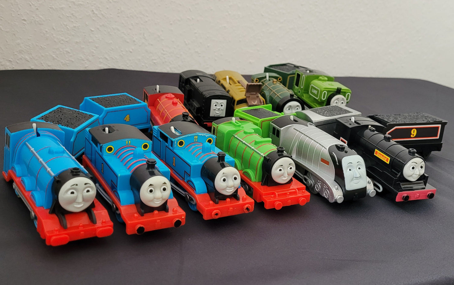 Das Bild zeigt die Vorderseite einer Sammlung von Thomas und seine Freunde-Zügen, die sorgfältig auf einem schwarzen Untergrund arrangiert sind. Im Hintergrund hebt sich ein weißer Hintergrund ab, wodurch die verschiedenen Lokomotiven besonders gut zur Geltung kommen.