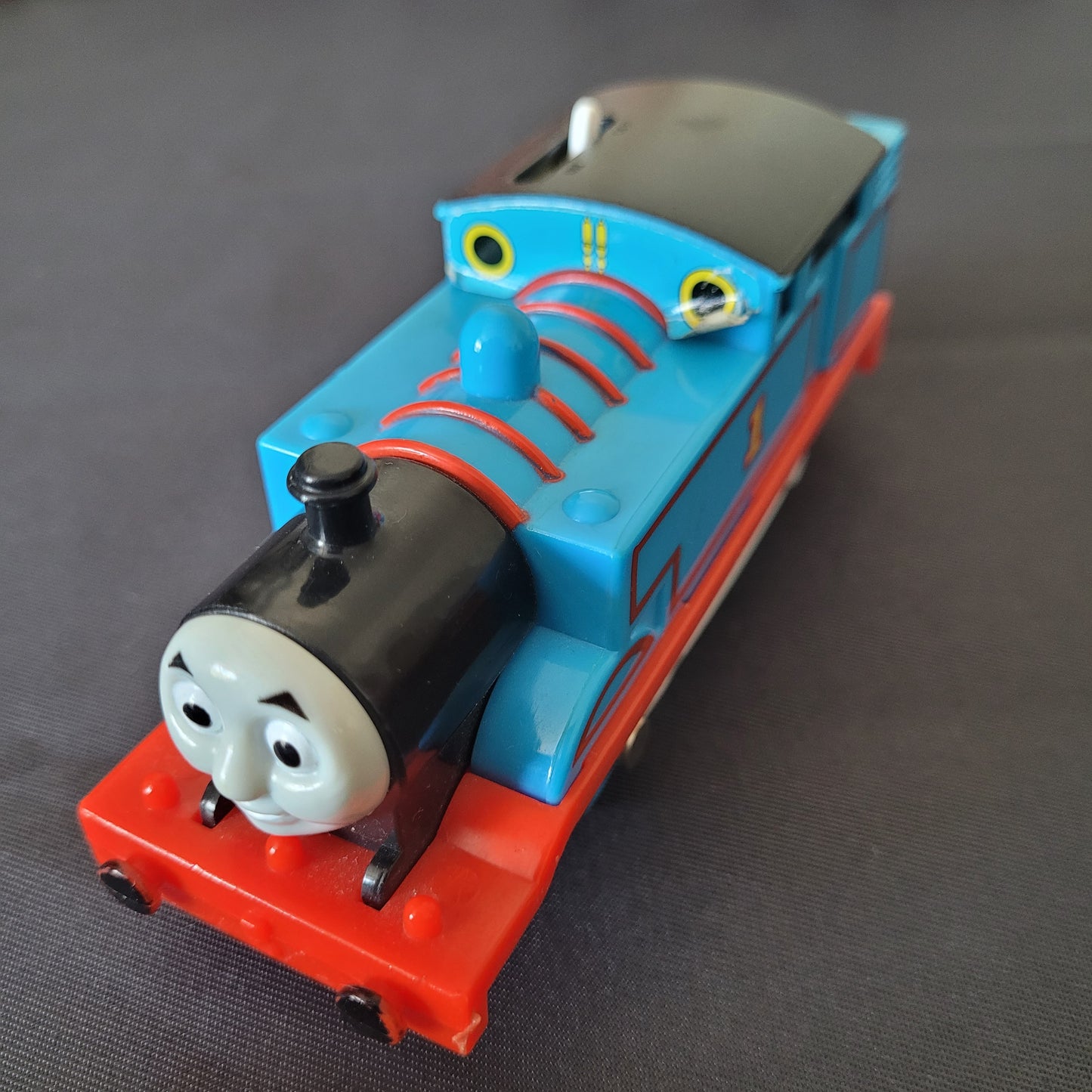 Das Bild zeigt die Lokomotive Thomas, Modell 1840D1, auf einem schwarzen Untergrund. Die blaue Lok mit rotem Fahrgestell und grauen Rädern ist detailliert gestaltet und zeigt die bekannten Gesichtszüge von Thomas aus der Serie ‚Thomas und seine Freunde‘