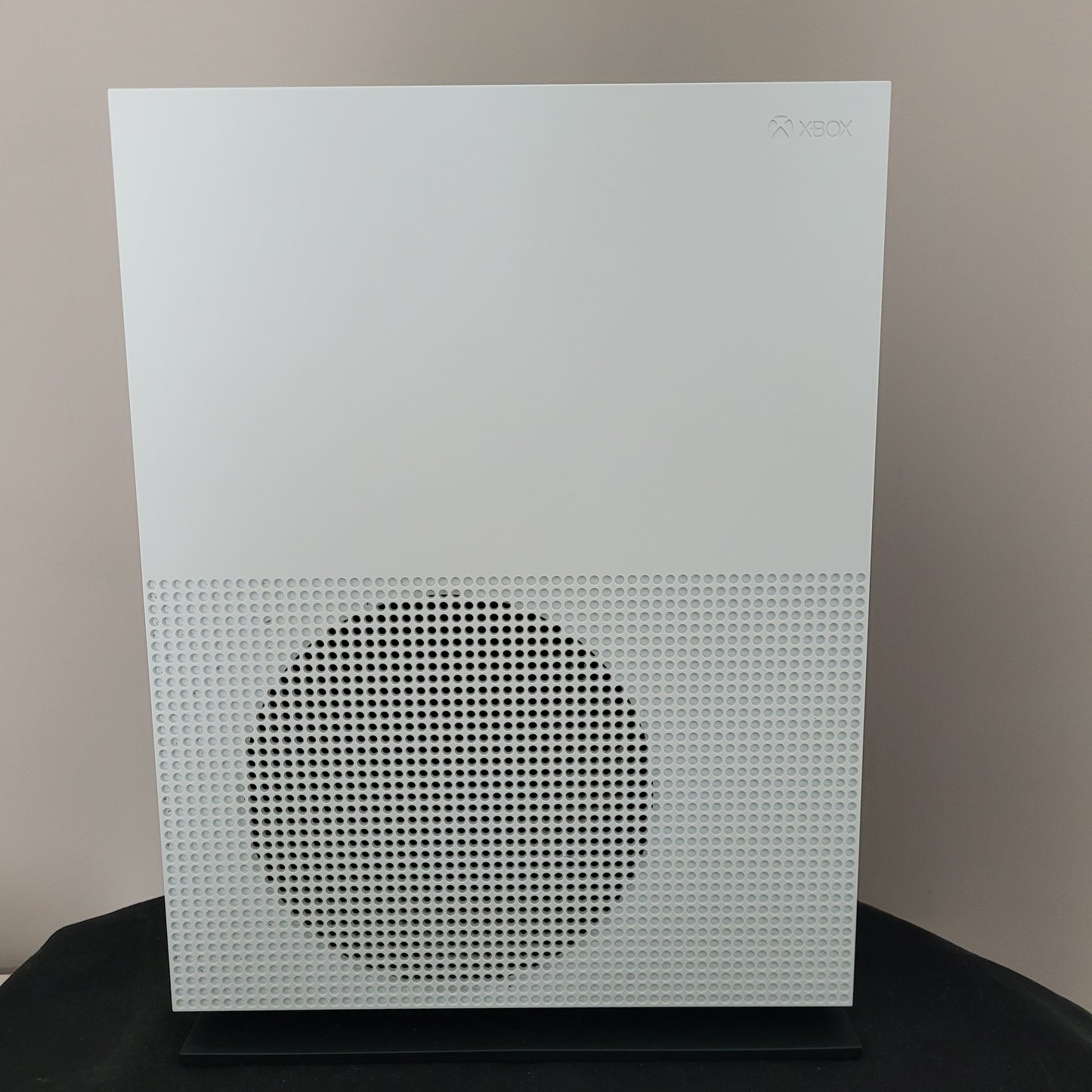 Das Foto zeigt die Seitenansicht einer weißen Xbox One S Konsole in vertikaler Position. Erkennbar ist das charakteristische große, runde Lüftungsgitter mit Wabenstruktur auf der rechten Geräteseite, während das dezente Xbox-Logo links oben eingelassen ist.