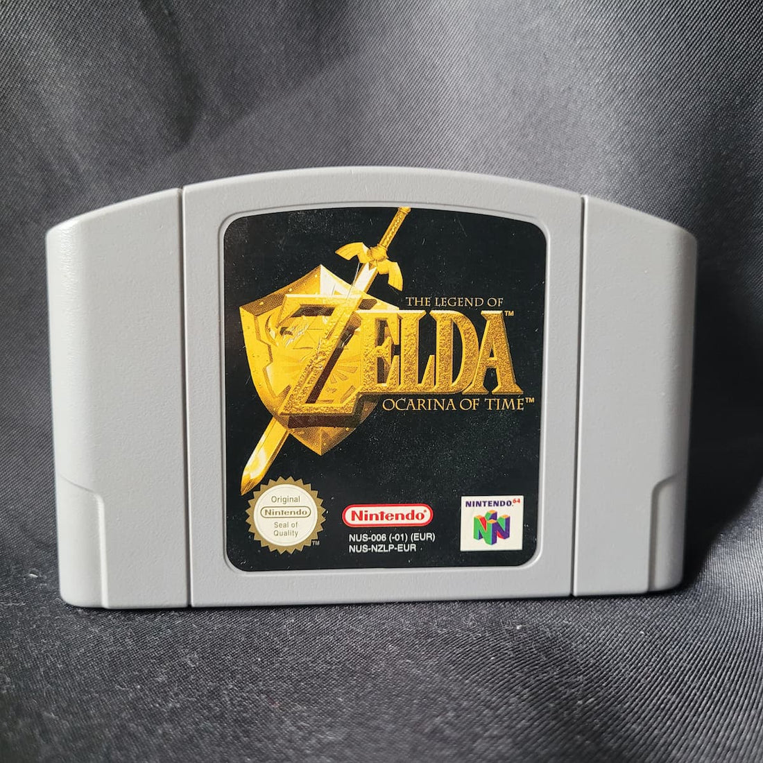 Nahaufnahme des grauen Nintendo 64 (N64) Spielemoduls von "The Legend of Zelda: Ocarina of Time" (PAL/EUR-Version) mit dem goldenen Logo auf schwarzem Hintergrund und dem Nintendo Seal of Quality.