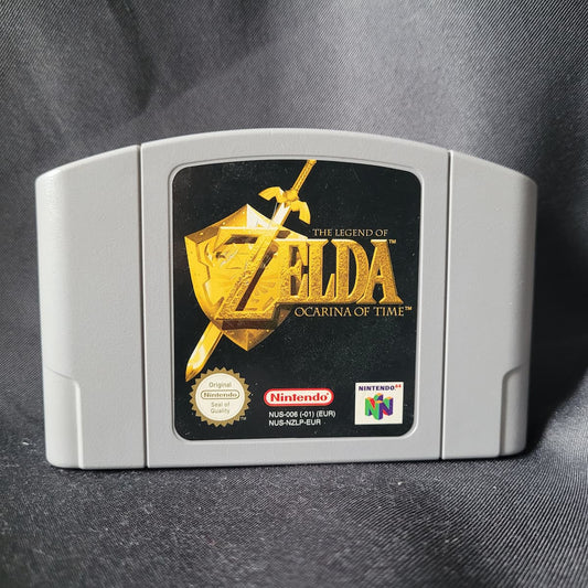 Nahaufnahme des grauen Nintendo 64 (N64) Spielemoduls von "The Legend of Zelda: Ocarina of Time" (PAL/EUR-Version) mit dem goldenen Logo auf schwarzem Hintergrund und dem Nintendo Seal of Quality.
