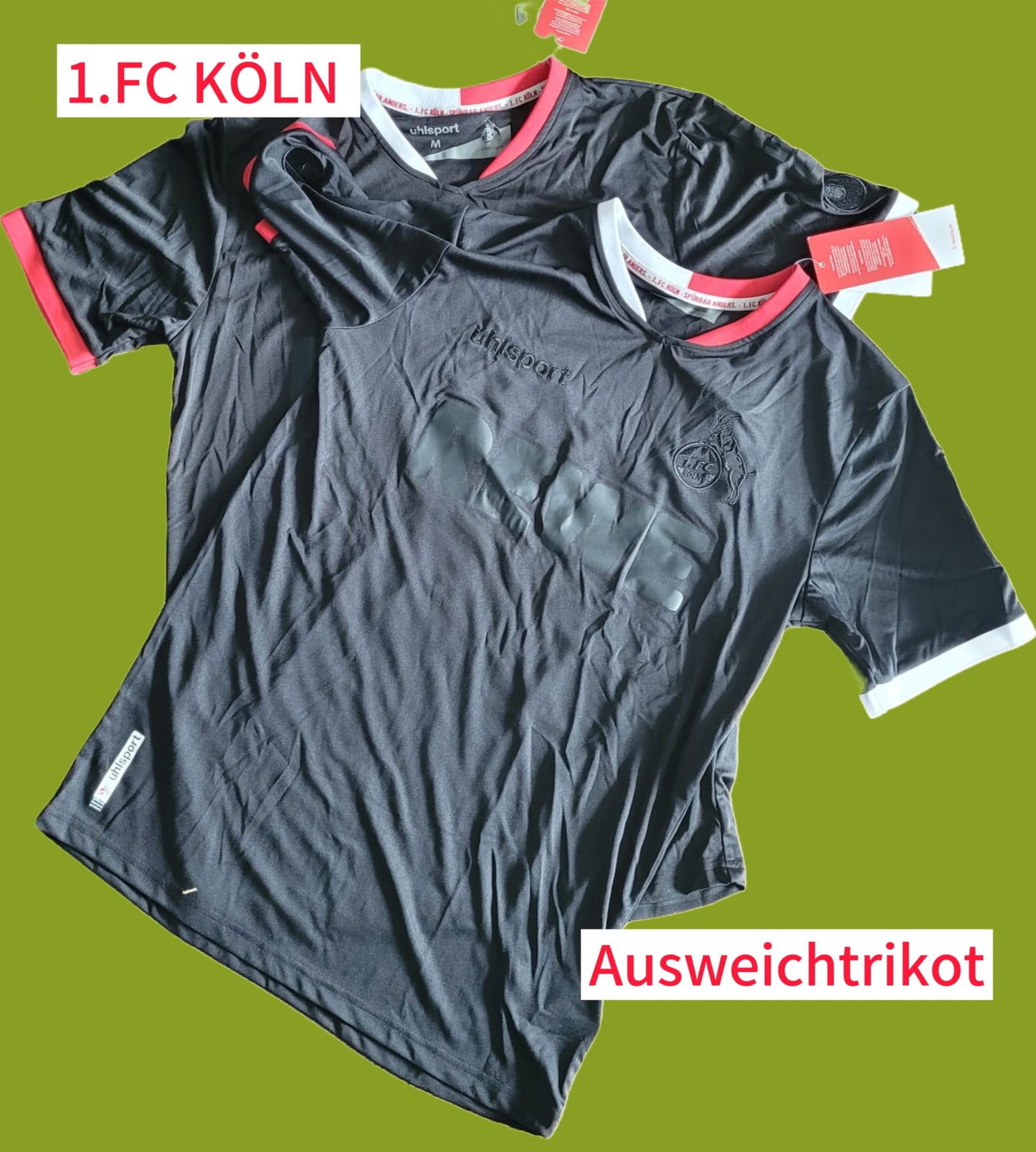 Zwei ungetragene Ausweichtrikots des 1. FC Köln aus der Saison 2021/22 liegen nebeneinander auf einem olivgrünen Untergrund. Beide Trikots sind schwarz mit roten und weißen Akzenten an Kragen und Ärmeln. In Größe M.