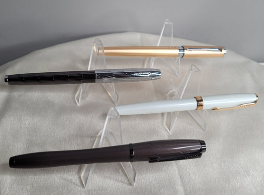 "Das Bild zeigt vier Füllfederhalter, die auf durchsichtigen Ständern präsentiert werden. Die Füller sind wie folgt beschrieben:
Parker Füllfederhalter metallic brown: Ein Füller mit einer metallisch braunen Oberfläche.
Parker Füllfederhalter Sonnet Gold: Ein Füller mit einer goldenen Oberfläche.
Parker Füllfederhalter Sonnet weiß-gold: Ein Füller mit einer weißen Oberfläche und goldenen Akzenten.
Parker Füllfederhalter Twin Metall Chiselled: Ein Füller mit einer metallischen, gravierten Oberfläche."