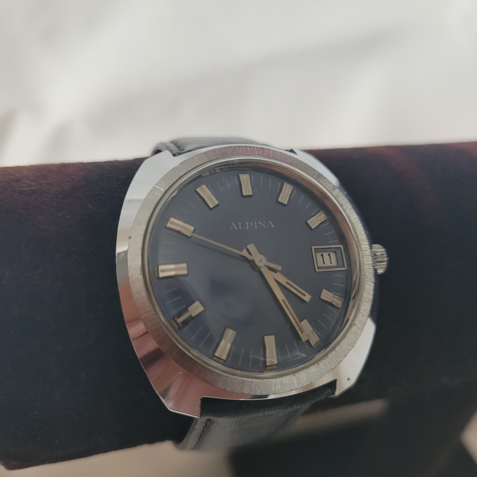 Auf dem Foto ist eine Alpina Vintage Armbanduhr zu sehen. Sie verfügt über ein rundes Edelstahlgehäuse, ein dunkles Zifferblatt mit rechteckigen Stundenmarkierungen sowie eine Datumsanzeige bei 3 Uhr. Die Uhr ist mit einem schwarzen Lederarmband ausgestattet, das den eleganten Charakter unterstreicht. Diese Alpina Vintage Uhr kombiniert klassisches Design mit Schweizer Präzision und ist ein begehrtes Sammlerstück für Liebhaber historischer Armbanduhren.