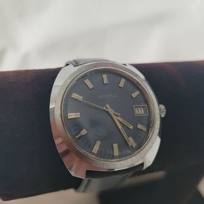 Auf dem Foto ist eine Alpina Vintage Armbanduhr zu sehen. Sie verfügt über ein rundes Edelstahlgehäuse, ein dunkles Zifferblatt mit rechteckigen Stundenmarkierungen sowie eine Datumsanzeige bei 3 Uhr. Die Uhr ist mit einem schwarzen Lederarmband ausgestattet, das den eleganten Charakter unterstreicht. Diese Alpina Vintage Uhr kombiniert klassisches Design mit Schweizer Präzision und ist ein begehrtes Sammlerstück für Liebhaber historischer Armbanduhren.