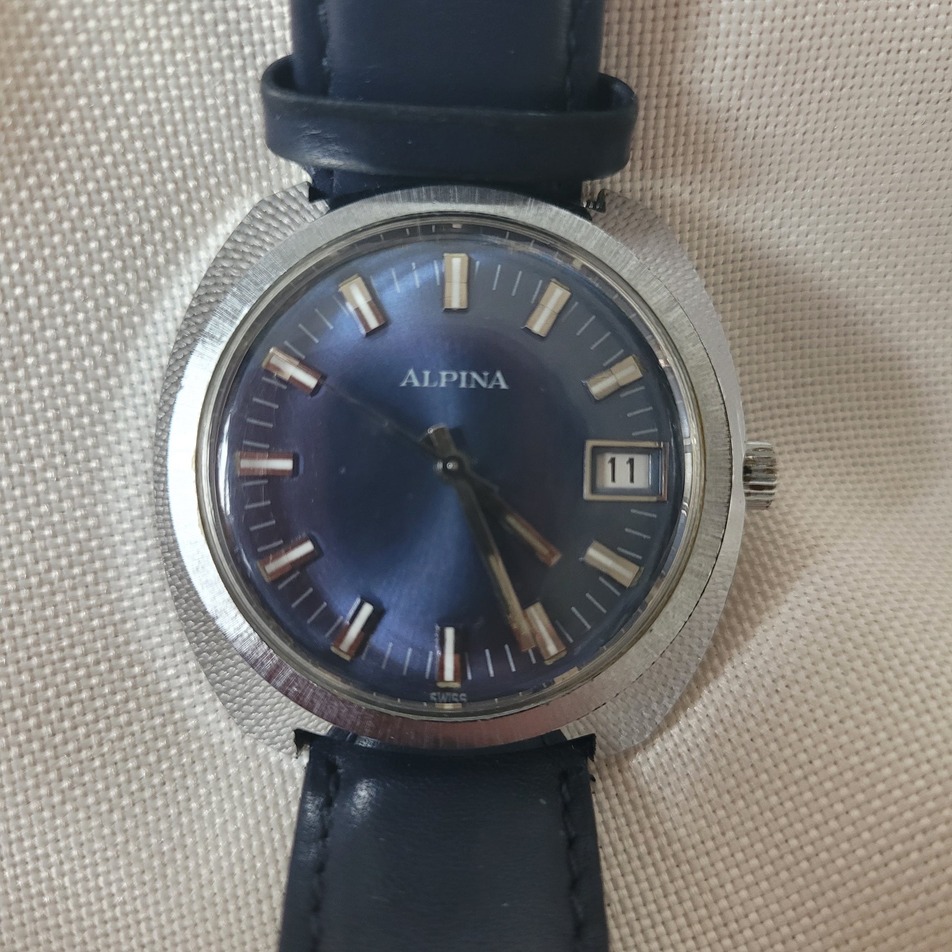 Alpina Vintage Armbanduhr mit blauem Zifferblatt, silbernem Edelstahlgehäuse und Datumsanzeige bei 12 Uhr. Die Uhr besitzt ein dunkelblaues Lederarmband und silberne Indizes mit Leuchtakzenten – klassische Schweizer Vintage-Uhr für Sammler.