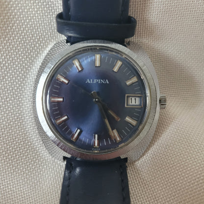 Alpina Vintage Armbanduhr mit blauem Zifferblatt, silbernem Edelstahlgehäuse und Datumsanzeige bei 12 Uhr. Die Uhr besitzt ein dunkelblaues Lederarmband und silberne Indizes mit Leuchtakzenten – klassische Schweizer Vintage-Uhr für Sammler.