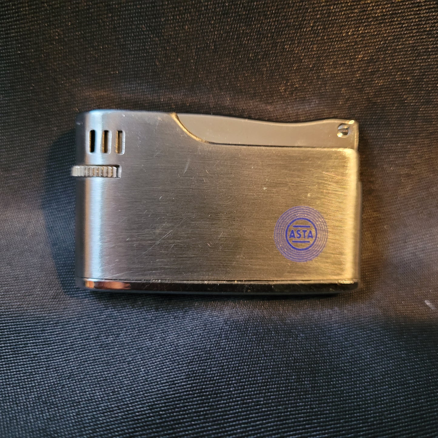 Apollo-K Feuerzeug Vintage, silberfarben, Vorderseite mit blau graviertem ASTA Logo, Hintergrund schwarz, kompaktes Sammlerstück 5,5 × 3 × 1 cm.