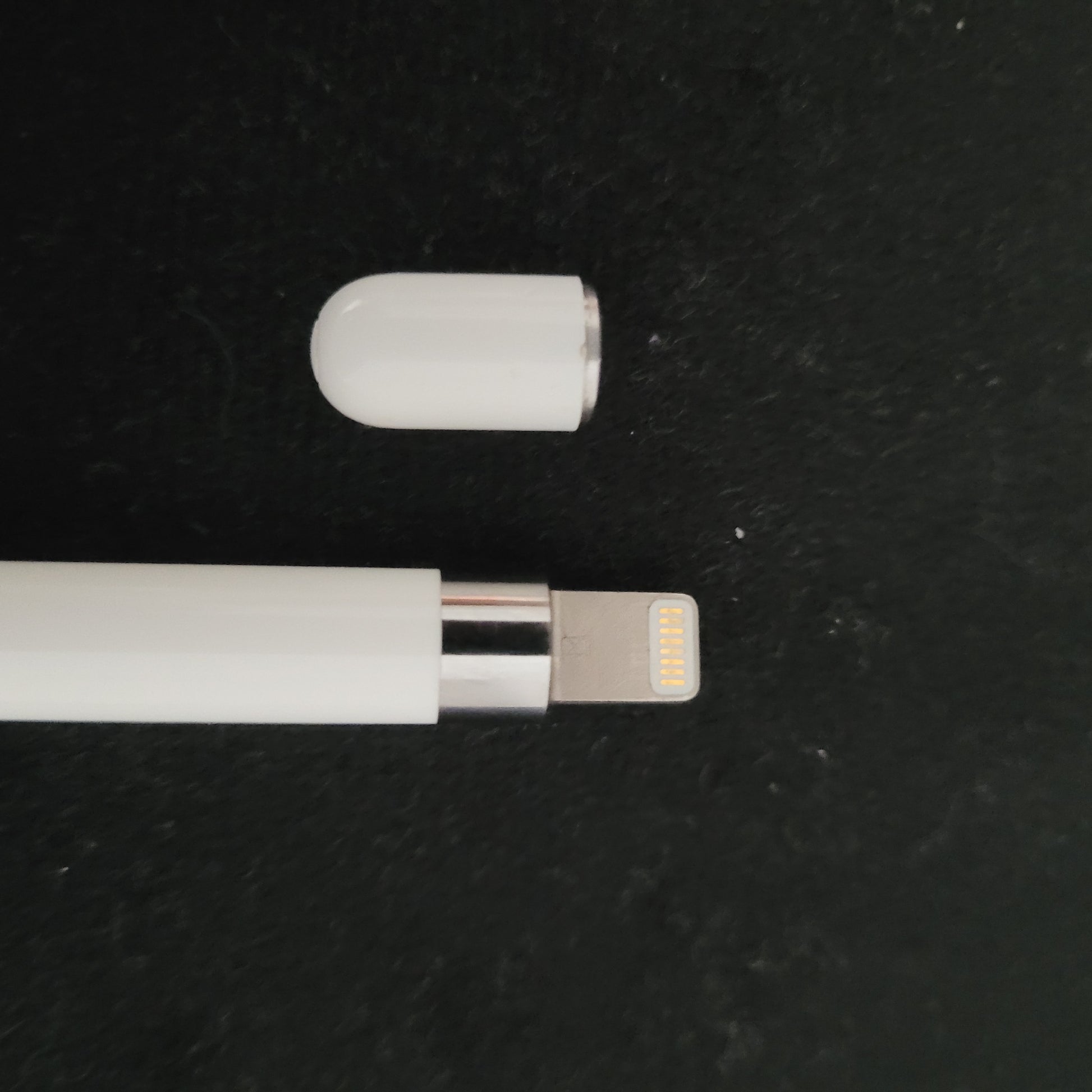 Ein weißer Apple Pencil mit abgenommener Kappe liegt auf einer schwarzen Oberfläche; der metallische Lightning-Anschluss ist sichtbar, die Kappe daneben platziert.