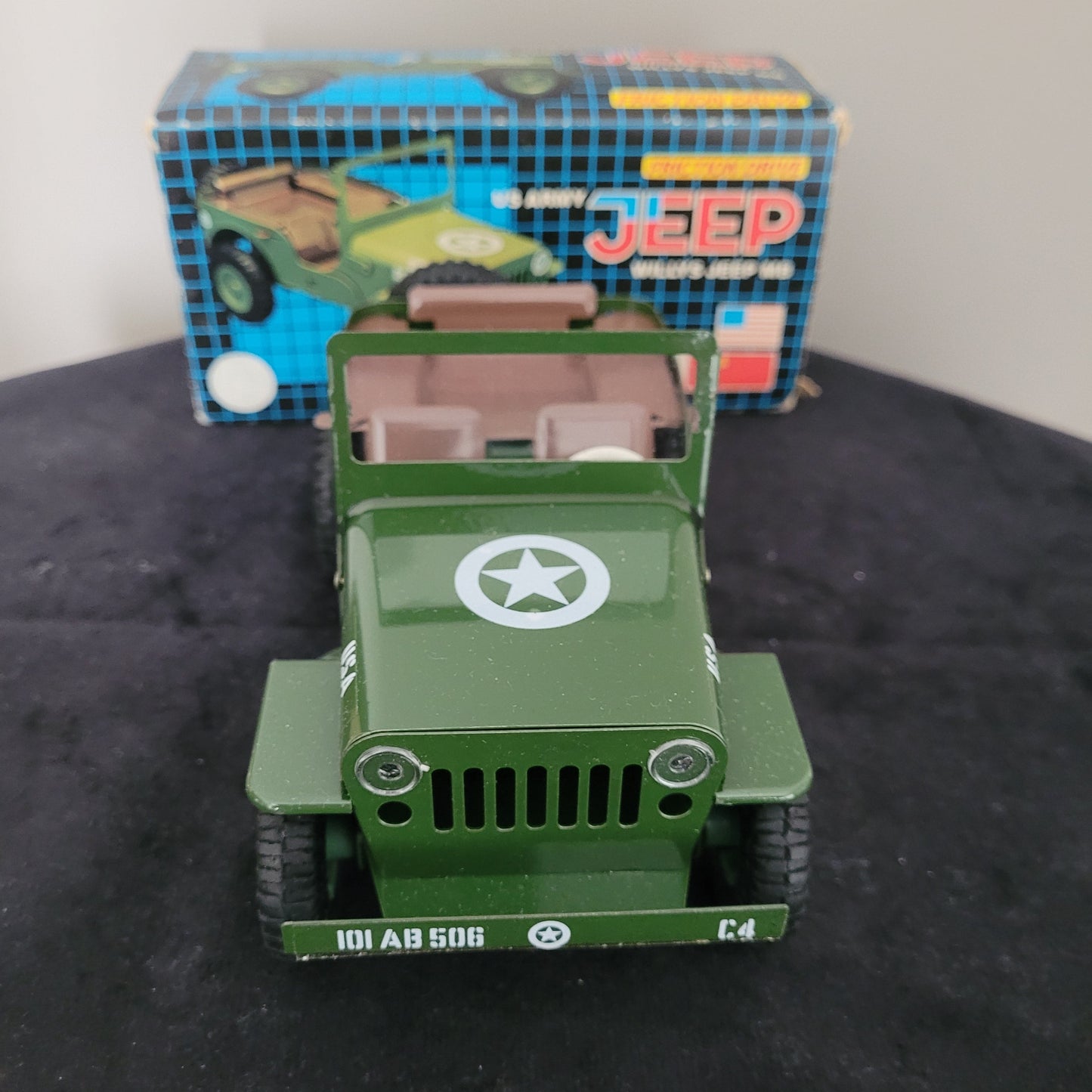 Das Bild zeigt die Vorderseite des Vintage-Blechspielzeugs Lotus Militär US Army Willys Jeep MB, sorgfältig platziert auf einem schwarzen Untergrund. Im Hintergrund sind ein weißer Bereich sowie die Originalverpackung (OVP) sichtbar, die das klassische Design des angegebenen Produkts unterstreichen