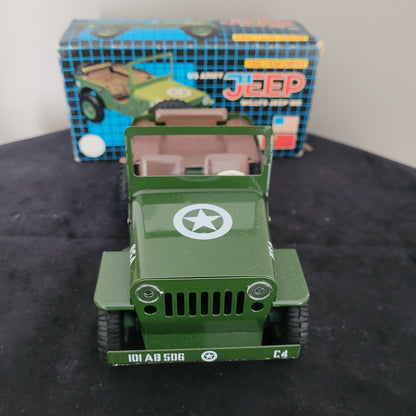 Das Bild zeigt die Vorderseite des Vintage-Blechspielzeugs Lotus Militär US Army Willys Jeep MB, sorgfältig platziert auf einem schwarzen Untergrund. Im Hintergrund sind ein weißer Bereich sowie die Originalverpackung (OVP) sichtbar, die das klassische Design des angegebenen Produkts unterstreichen