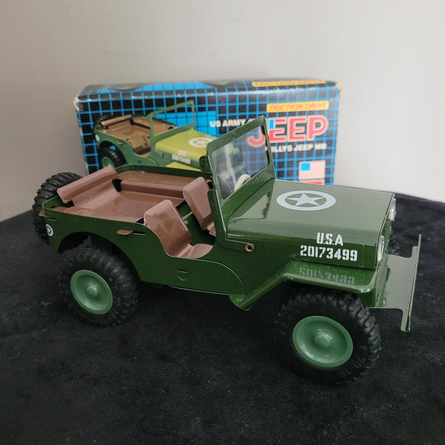 Das Bild zeigt die Vorderseite des Vintage-Blechspielzeugs Lotus Militär US Army Willys Jeep MB, sorgfältig seitlich platziert auf einem schwarzen Untergrund. Im Hintergrund sind ein weißer Bereich sowie die Originalverpackung (OVP) sichtbar, die das klassische Design des angegebenen Produkts unterstreichen