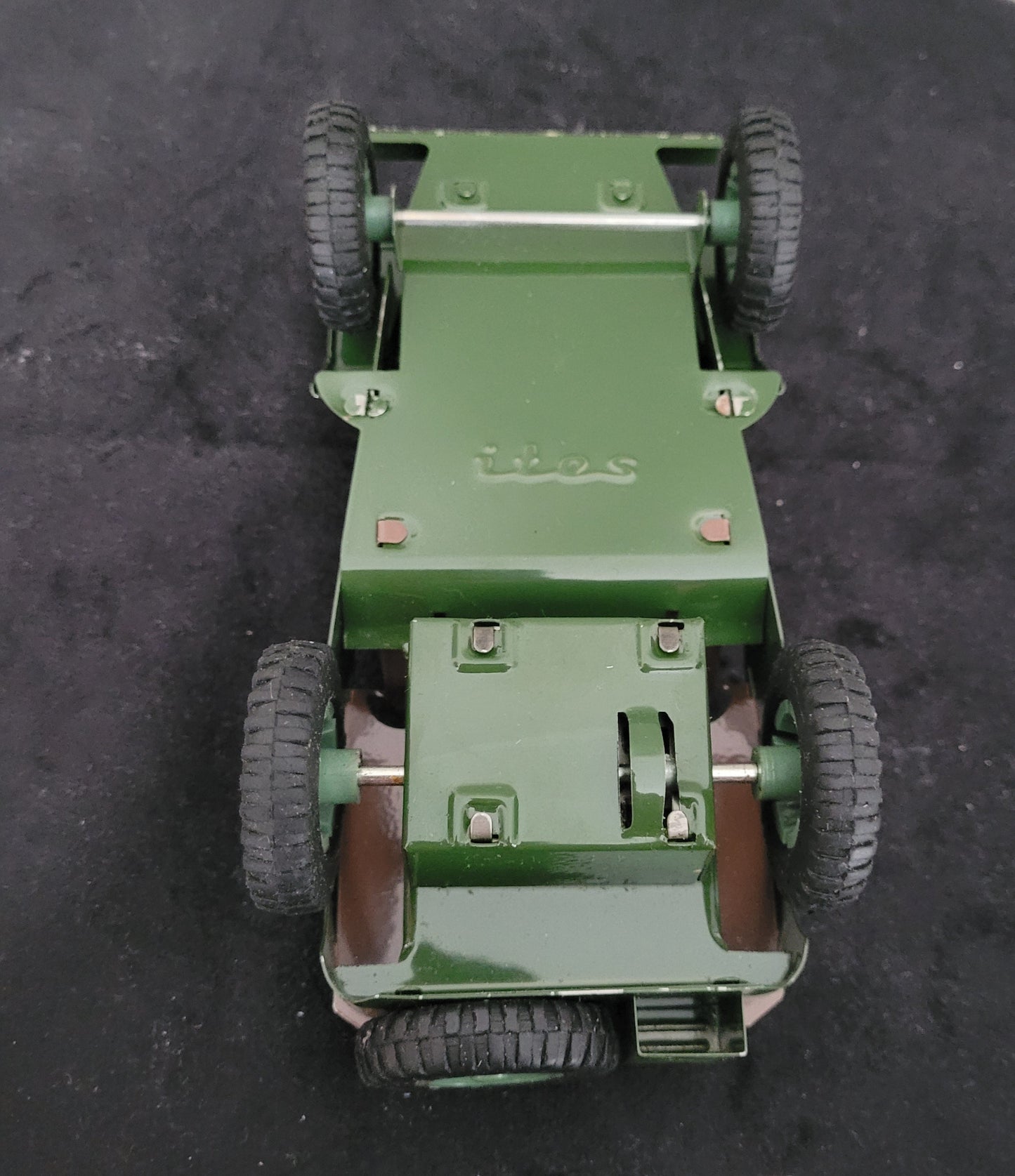 Das Bild zeigt die Unterseite des Vintage-Blechspielzeugs Lotus Militär US Army Willys Jeep MB, platziert auf einem schwarzen Untergrund. Sichtbar sind die mechanischen Details, Räder und die robuste Blechkonstruktion, die den klassischen Retro-Charme des angegebenen Produkts unterstreichen.