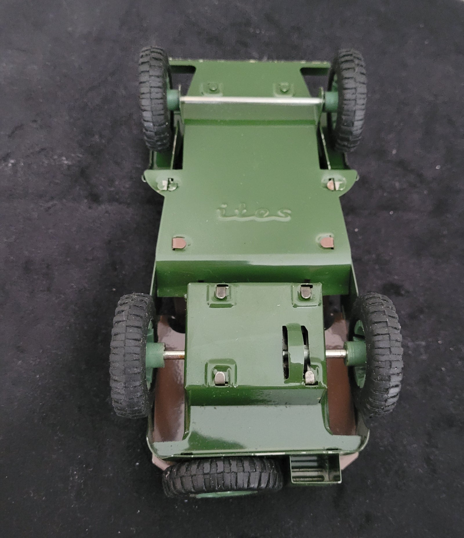 Das Bild zeigt die Unterseite des Vintage-Blechspielzeugs Lotus Militär US Army Willys Jeep MB, platziert auf einem schwarzen Untergrund. Sichtbar sind die mechanischen Details, Räder und die robuste Blechkonstruktion, die den klassischen Retro-Charme des angegebenen Produkts unterstreichen.