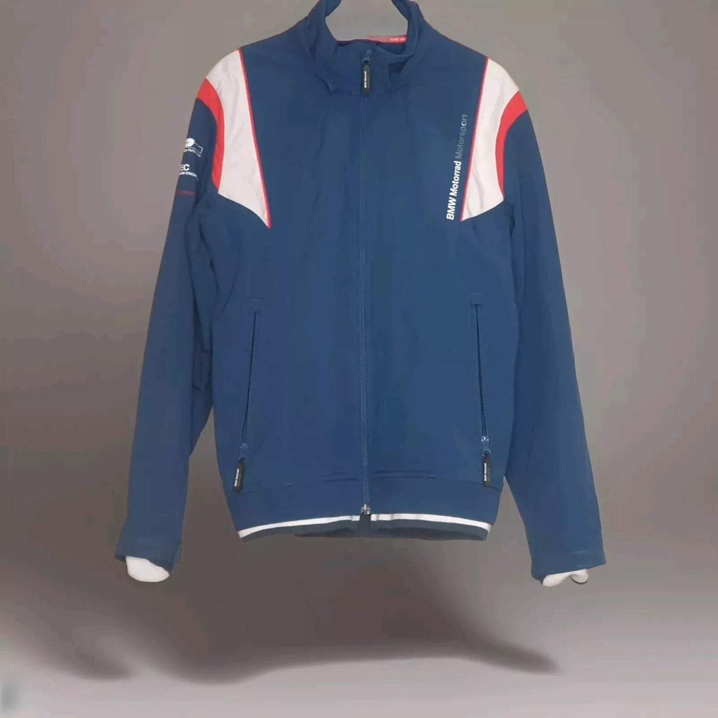 Blaue BMW Motorrad Motorsport Jacke mit Reißverschluss und weißen sowie roten Akzenten auf den Schultern, aufgehängt an einem Kleiderbügel. Ideal für Motorsport-Enthusiasten und Fans von BMW Motorrad Kleidung.