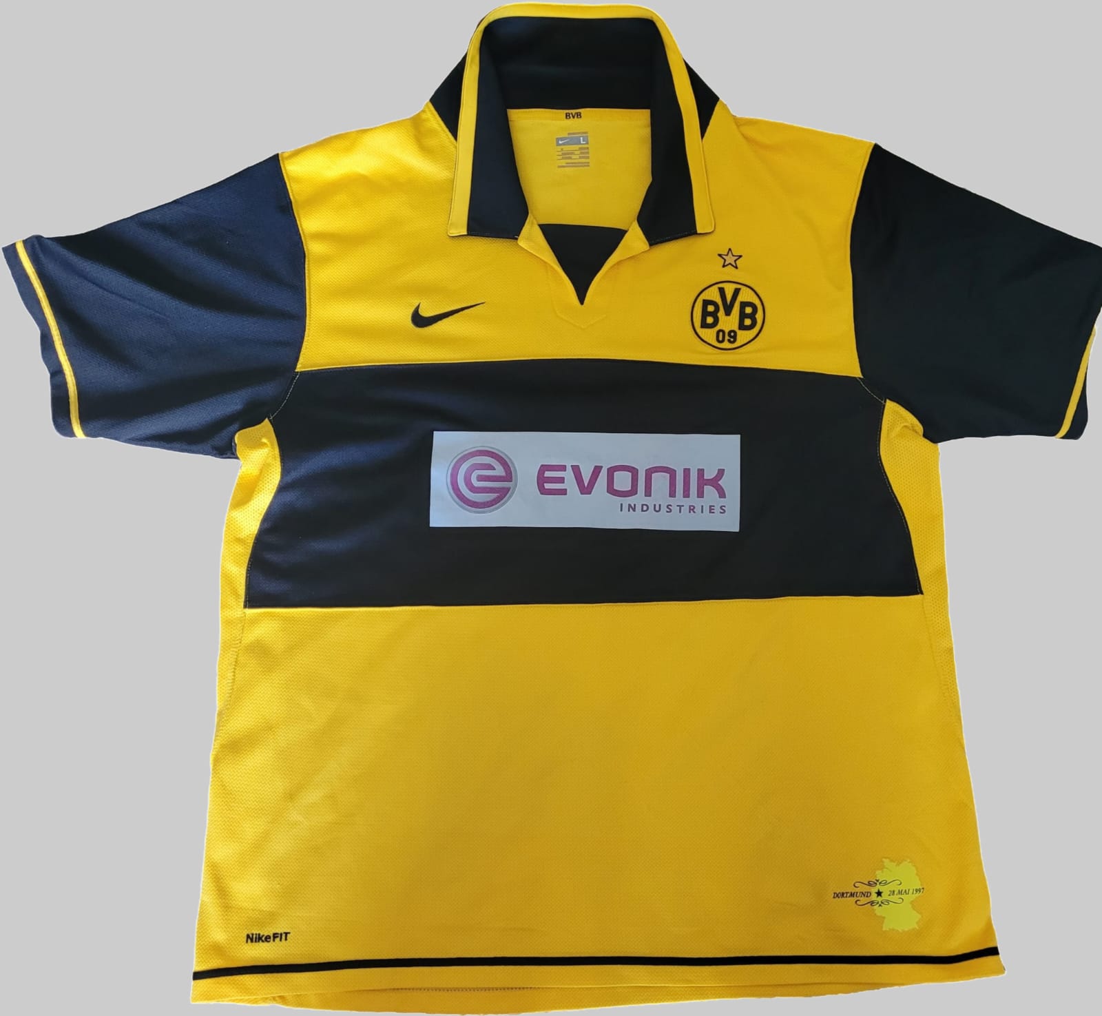 Borussia Dortmund BVB Vintage Trikot 2007-2008 – Vorderseite auf grauem Hintergrund Das Bild zeigt die Vorderseite eines Borussia Dortmund BVB Vintage Trikots aus der Saison 2007-2008, platziert auf einem grauen Hintergrund. Das Trikot ist überwiegend gelb mit schwarzen Akzenten an den Ärmeln und Seiten. Auf der linken Brustseite befindet sich das BVB-Logo mit einem Stern darüber, während auf der rechten Brustseite das Nike-Logo sichtbar ist. In der Mitte des Trikots prangt das Logo des Sponsors „Evonik"